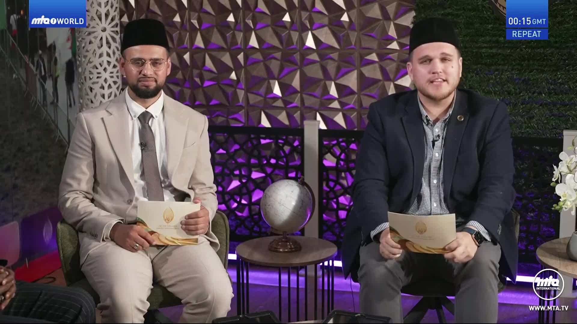 Two men in suits and traditional caps are seated and holding papers, likely prepared for a discussion on MTA1 World. A globe sits between them, and the backdrop features a geometric design.
