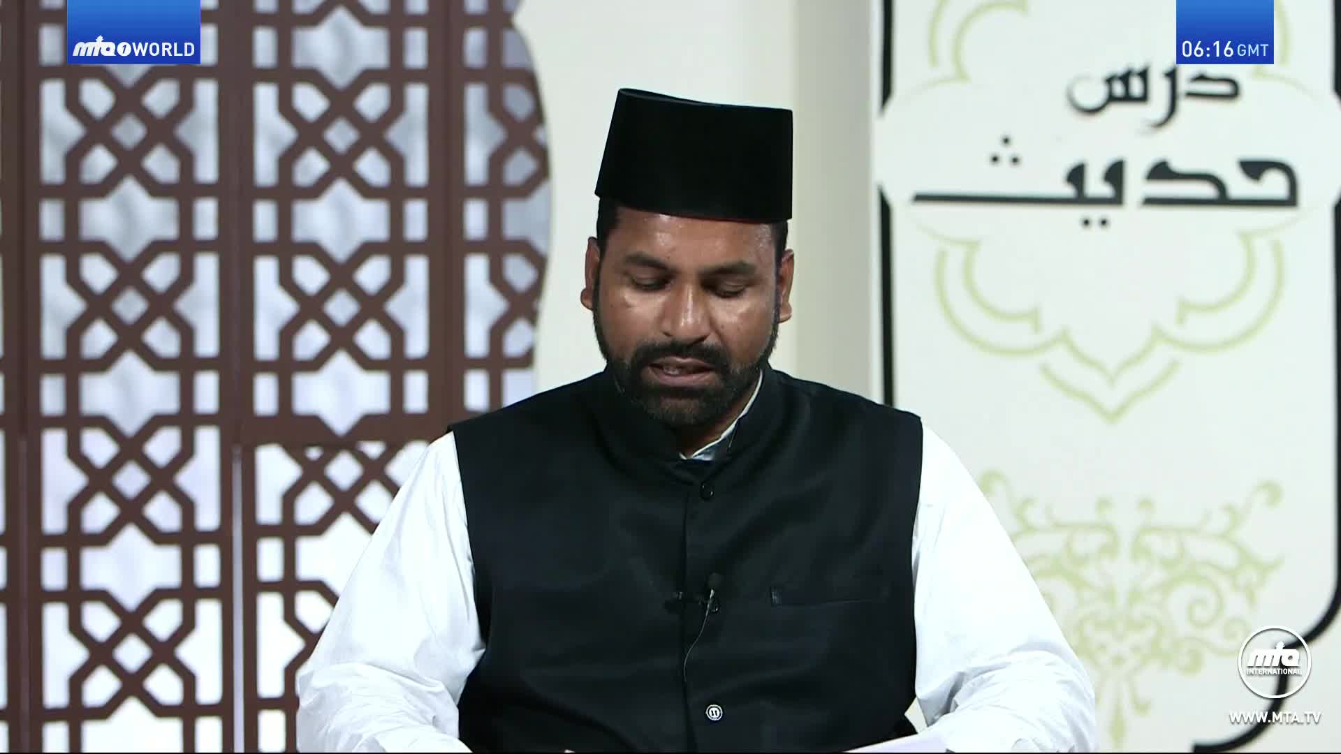 A man in a black cap and vest is speaking, his gaze focused downwards. Behind him, a decorative screen and a sign in Arabic script are visible.
