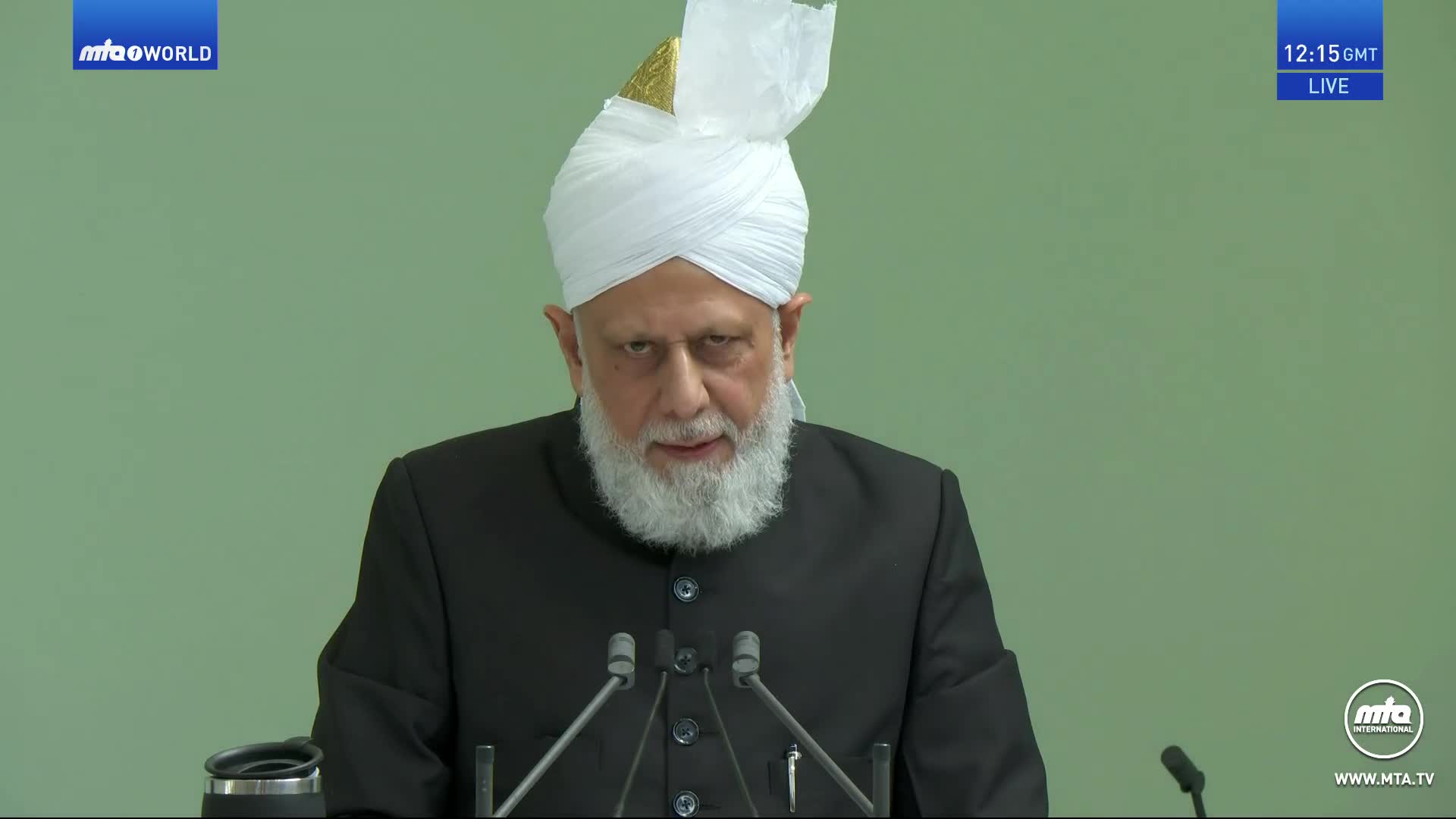 The man in the white turban is speaking, his expression serious as he addresses the microphones. The MTA1 World logo is visible in the corner, indicating a live broadcast from the United Kingdom.
