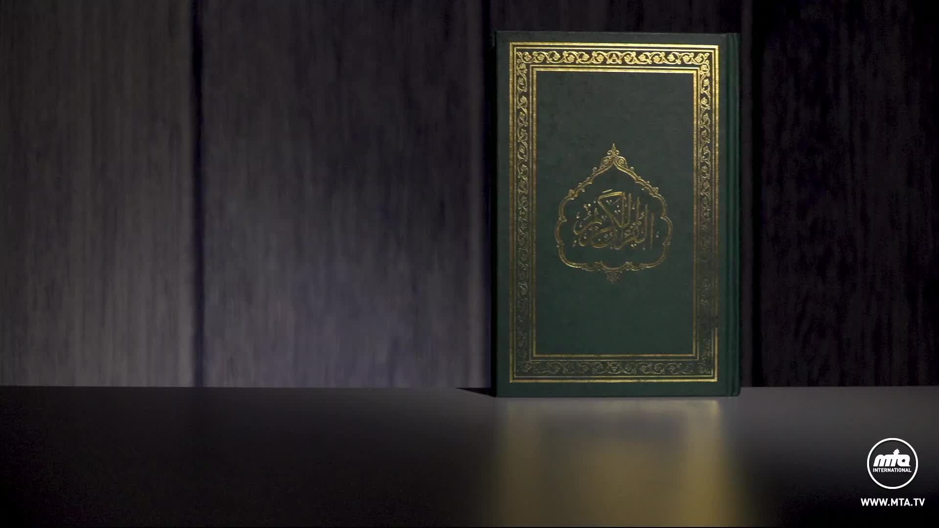 A green book with gold lettering sits upright on a dark surface. The cover features ornate calligraphy within a decorative border.
