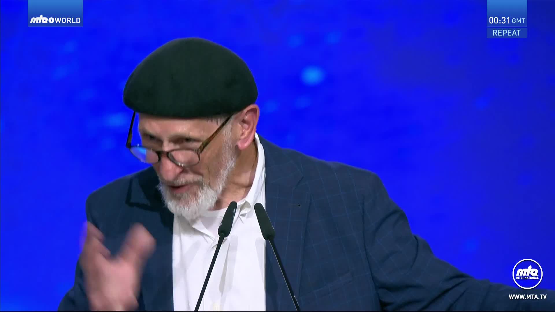 A man with a white beard and glasses gestures with his right hand, speaking into a microphone. He wears a dark green cap and a blue blazer, with the MTA World logo visible in the corner.
