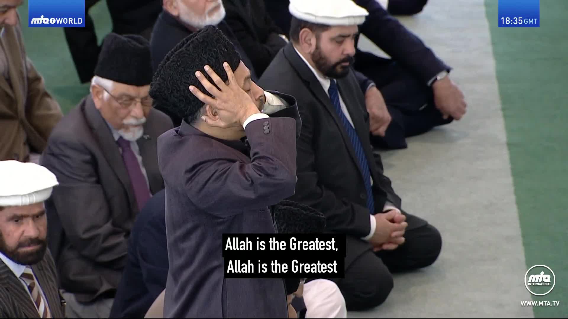 A man in a dark suit and a black hat raises his hands to his ears, palms forward, as he kneels. Other men, also in suits, sit nearby, participating in the prayer broadcast on MTA1 World from the United Kingdom.
