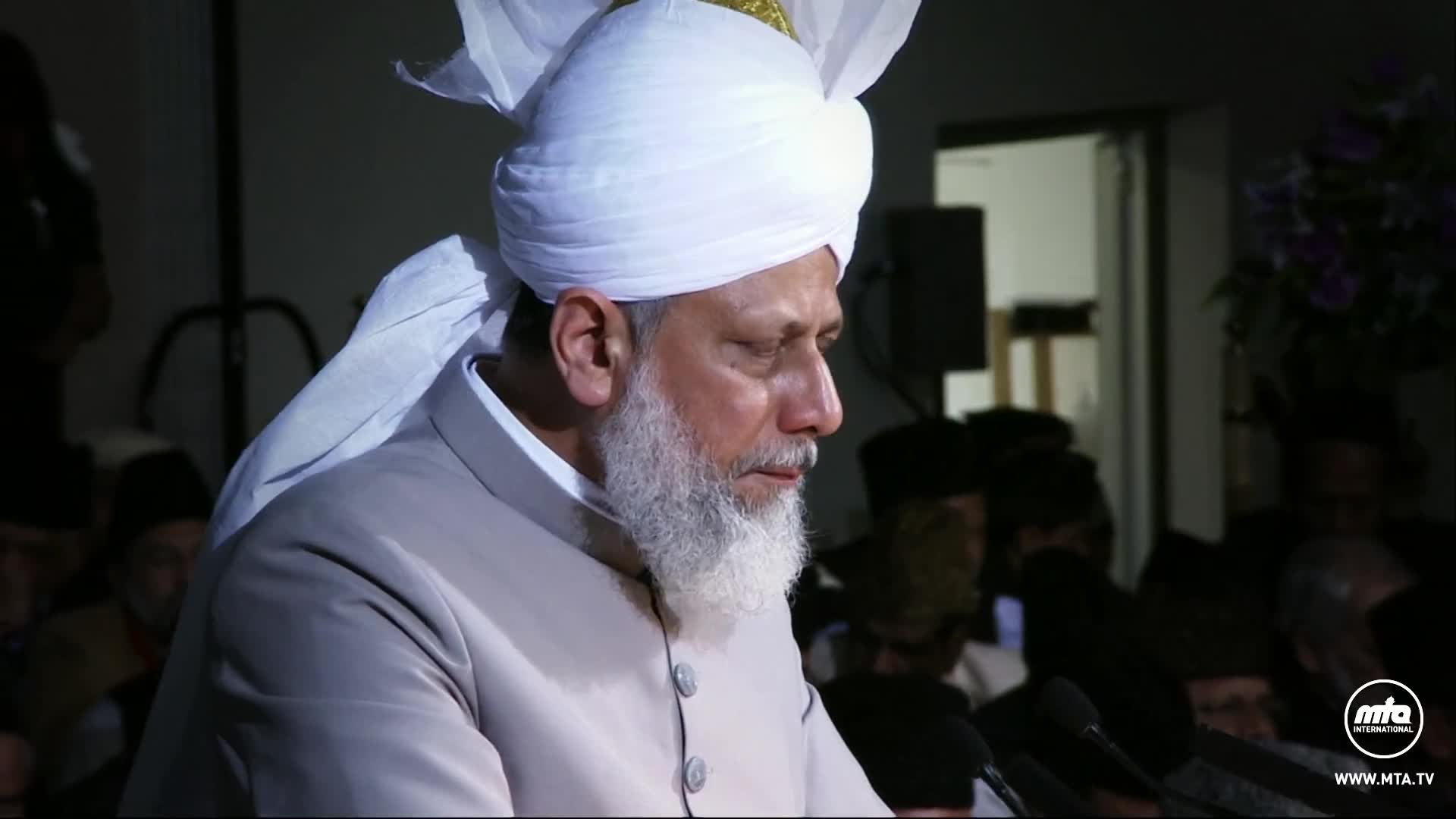 A man in a white turban and light-colored suit stands at a podium, speaking. His gaze is lowered, and a microphone is positioned in front of him.
