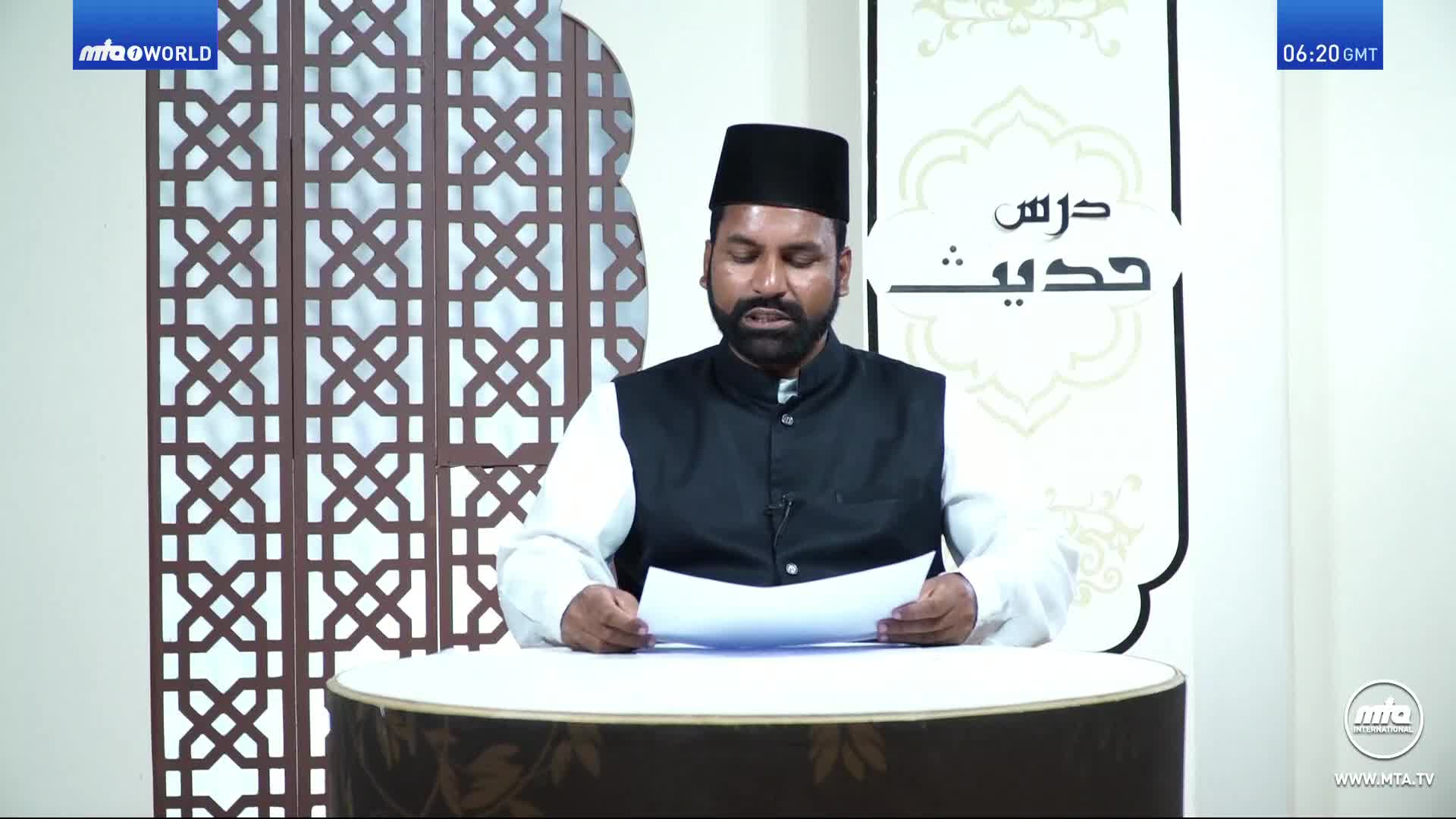 A man in a black cap and vest reads from papers at a desk, likely on MTA1 World. The backdrop features a decorative screen and Arabic script.

