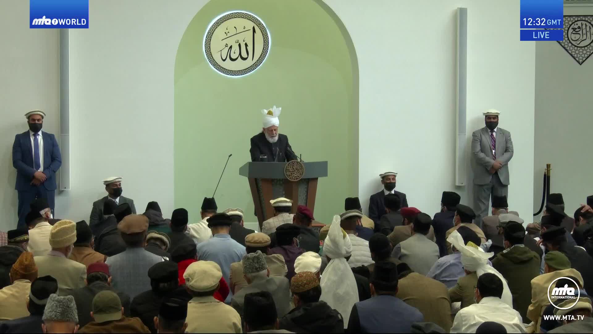A man in a white turban speaks from a podium, facing a crowd of people in the United Kingdom. Two men in suits stand attentively on either side of the speaker.

