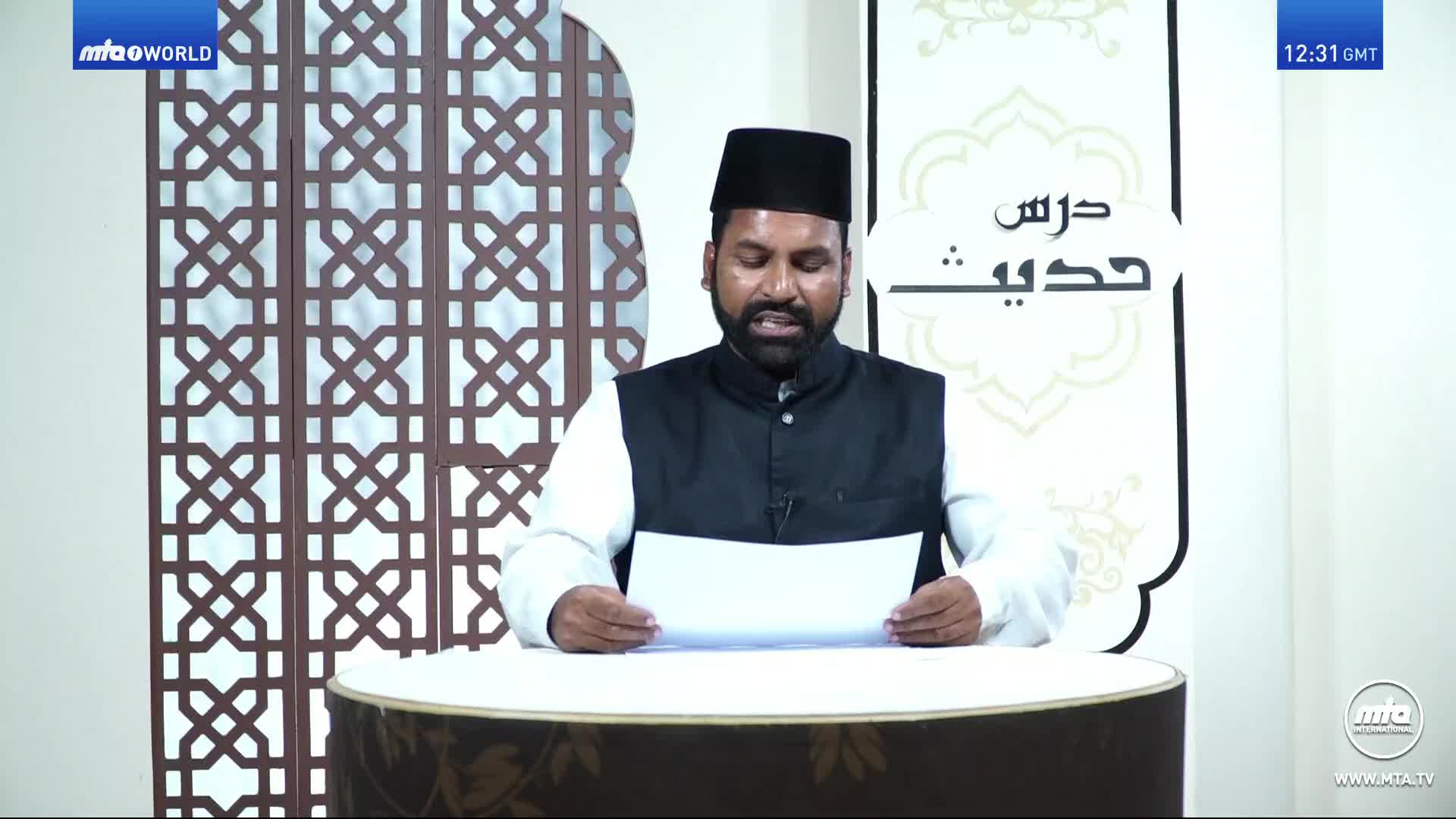 A man in a black cap and vest reads from a paper, his voice audible in the MTA1 World studio. Behind him, a decorative panel and a large banner with Arabic script provide the backdrop.
