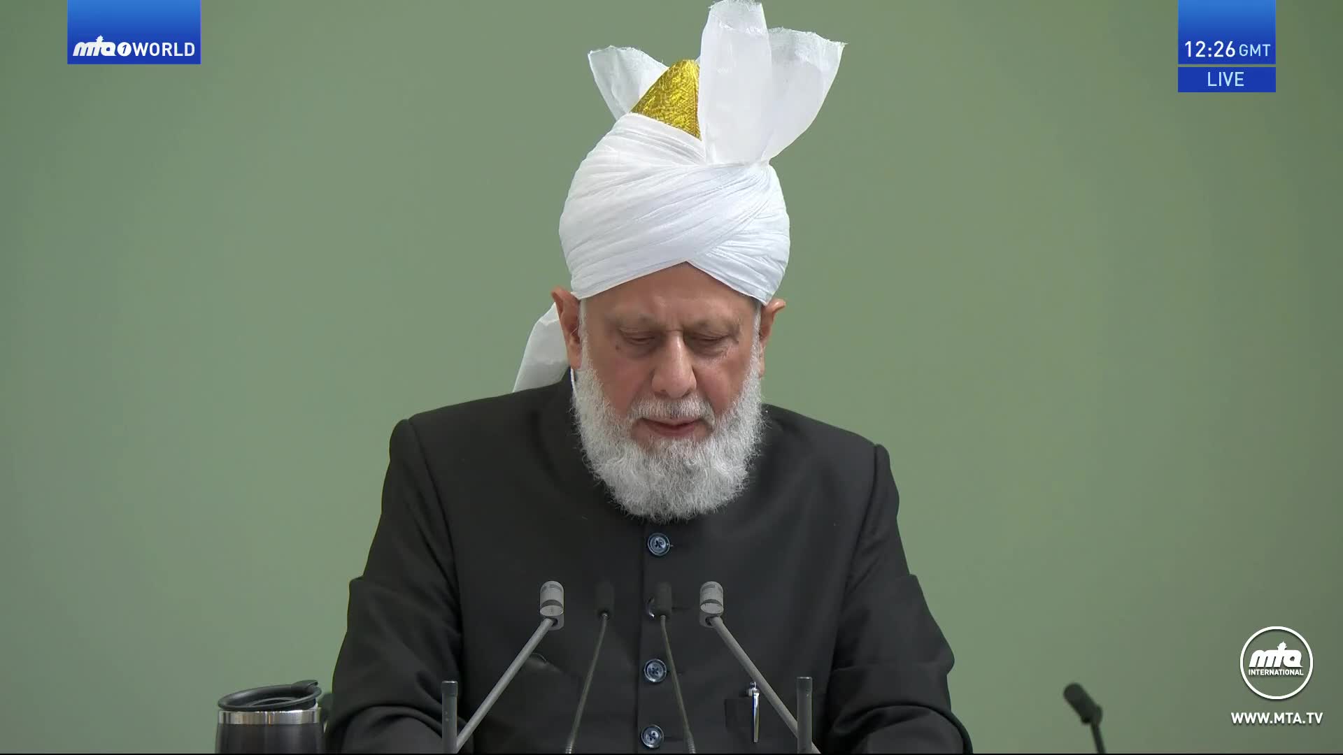 A man in a white turban and black suit speaks into several microphones. The MTA1 World logo is visible in the corner, and the time is displayed in the upper right.
