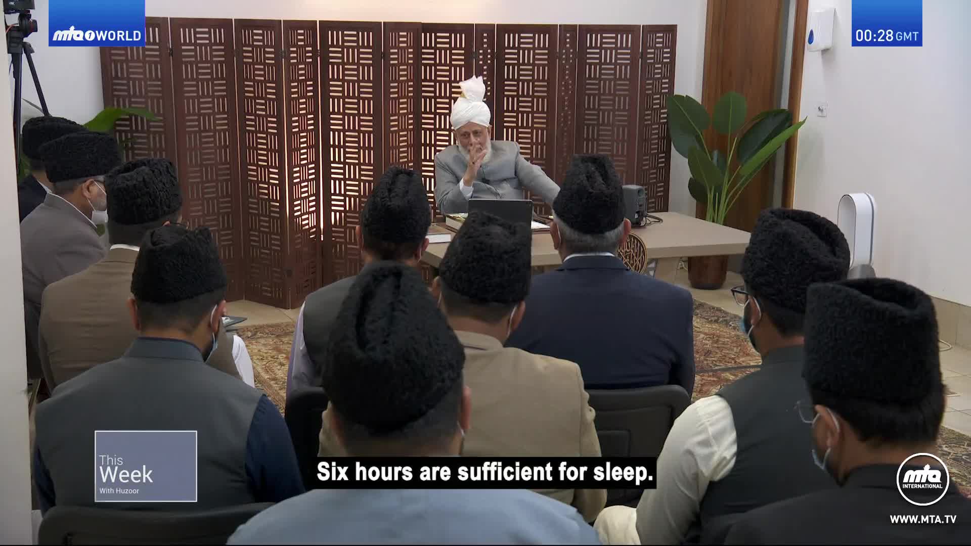 A man in a turban sits at a table, speaking to a group of men wearing black hats. The broadcast from MTA1 World in the United Kingdom is underway, with a caption about sleep appearing on the screen.
