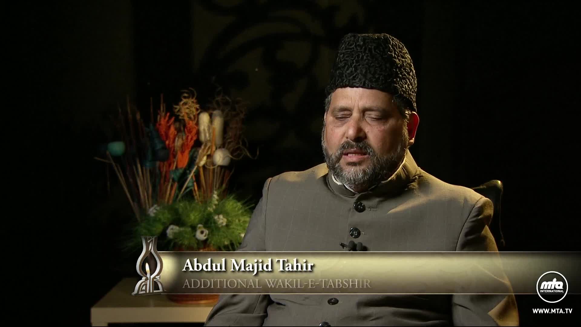 Abdul Majid Tahir, identified by the lower third, is speaking on camera. He wears a black cap and a grey suit, while a floral arrangement sits to his left.
