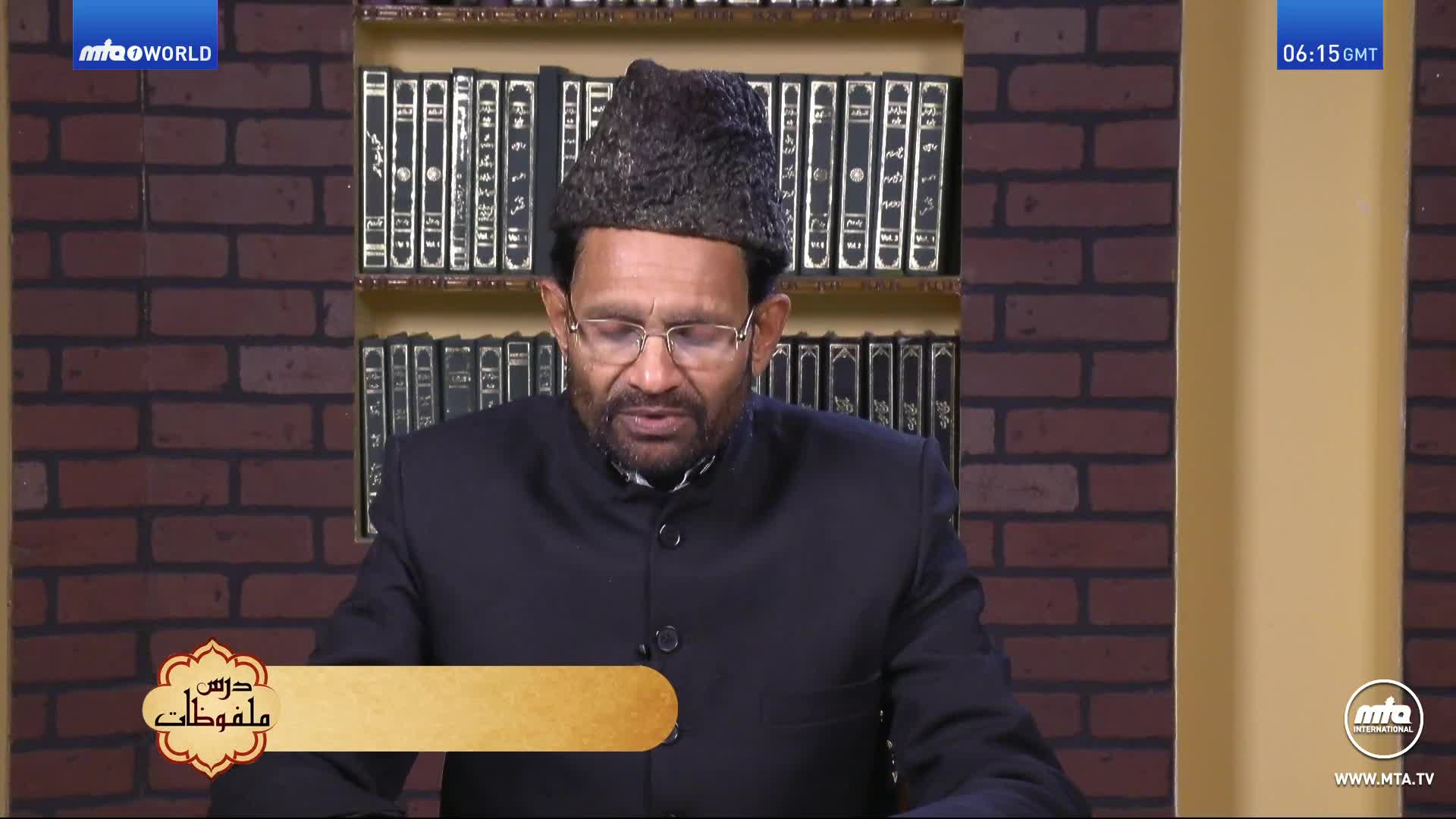 A man in a dark suit and hat is speaking, his gaze directed downwards. Behind him, a shelf is filled with books, and the MTA1 World logo is visible in the corner.
