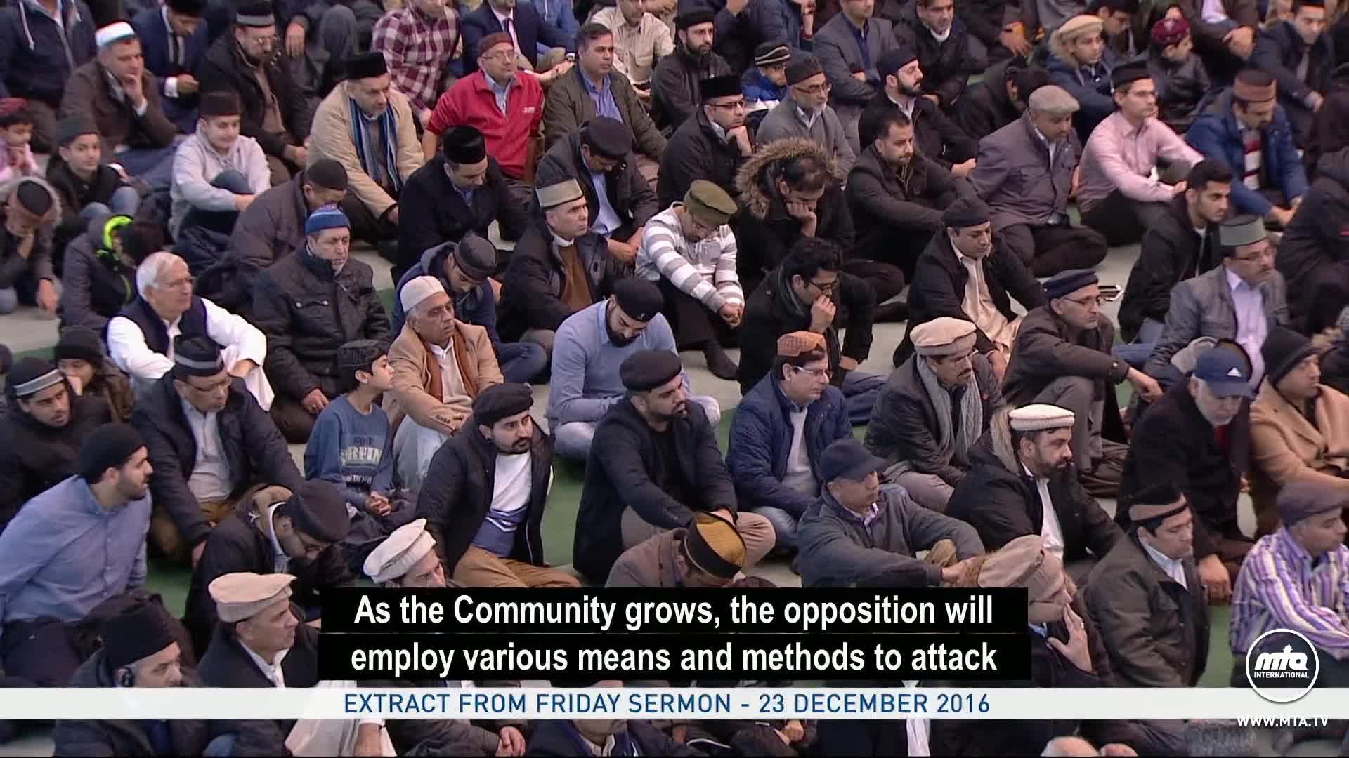 A large crowd of men are seated on the floor, many wearing hats and jackets, listening intently. The screen displays the MTA International logo and the date of a Friday sermon.

