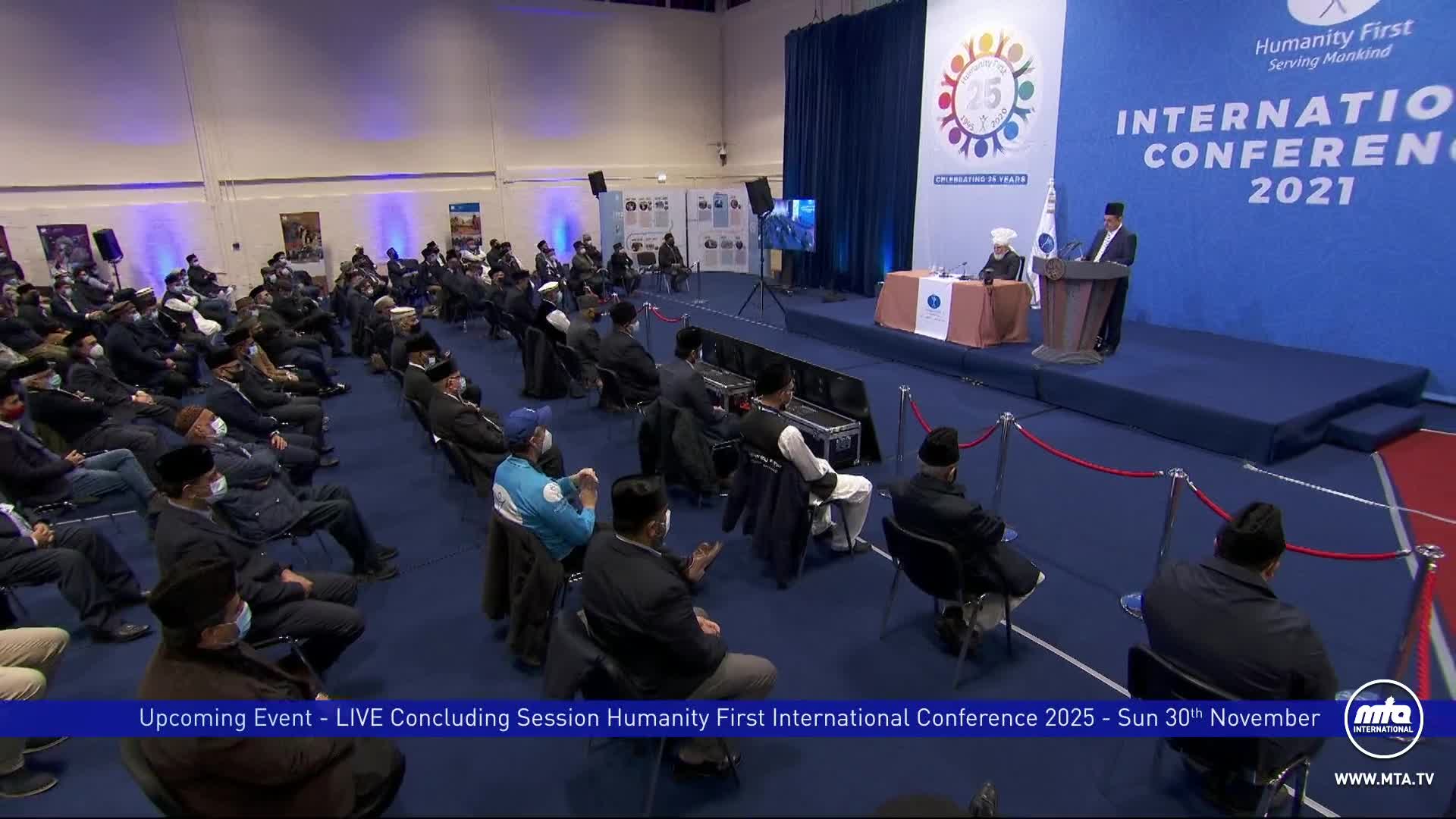 A man in a suit speaks from a podium, addressing a seated audience in a large hall. Most people are wearing masks, and the event is being broadcast live on MTA International.
