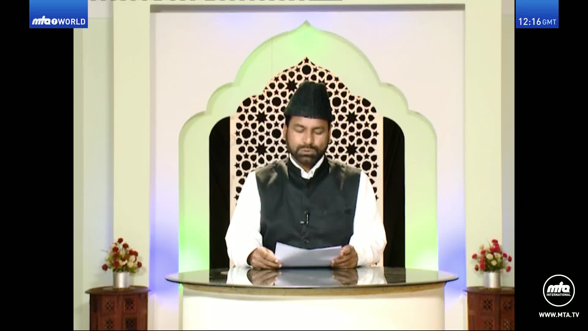 A man in a black cap and vest reads from a paper at a desk, the MTA World logo visible in the corner. Behind him, a decorative archway with a star pattern frames the backdrop.
