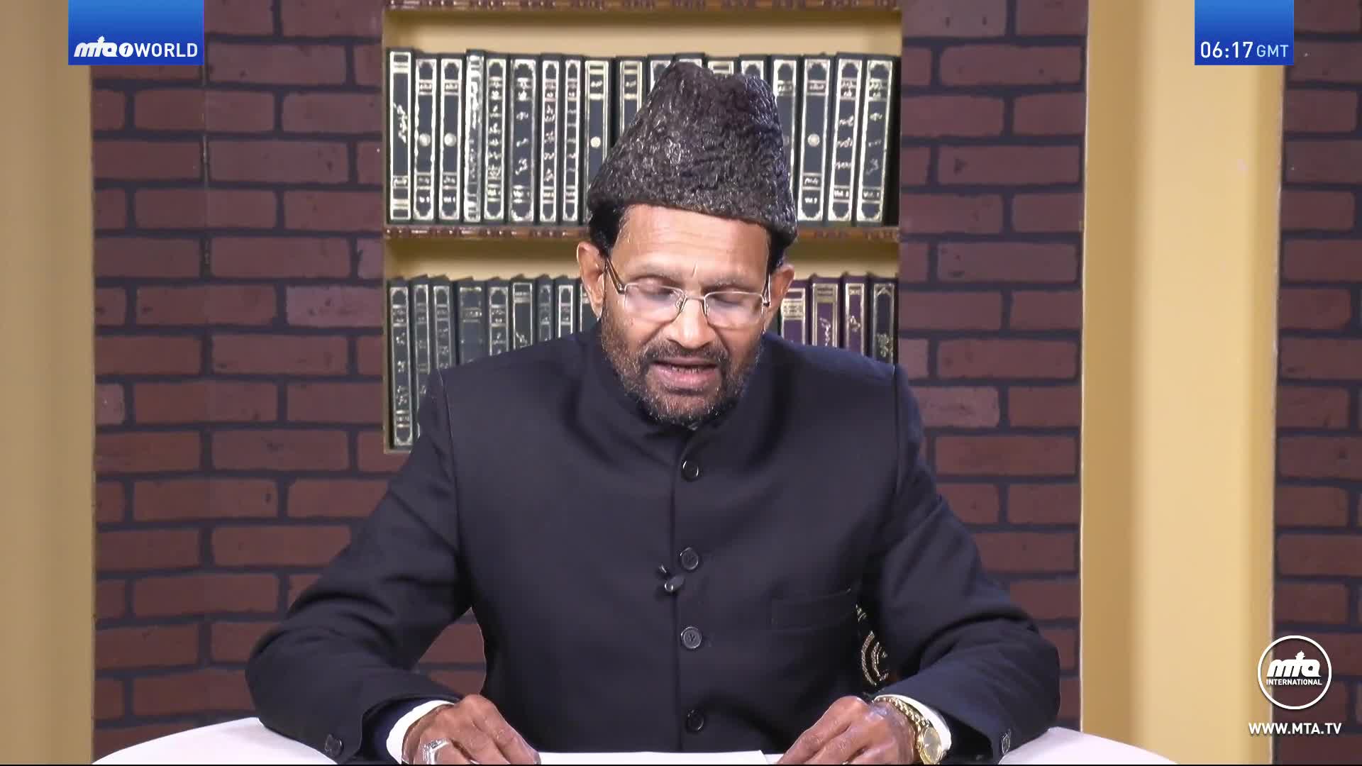 A man in a dark suit and a traditional hat speaks, his hands resting on papers in front of him. Behind him, a shelf is filled with books, and the MTA1 World logo is visible in the corner.
