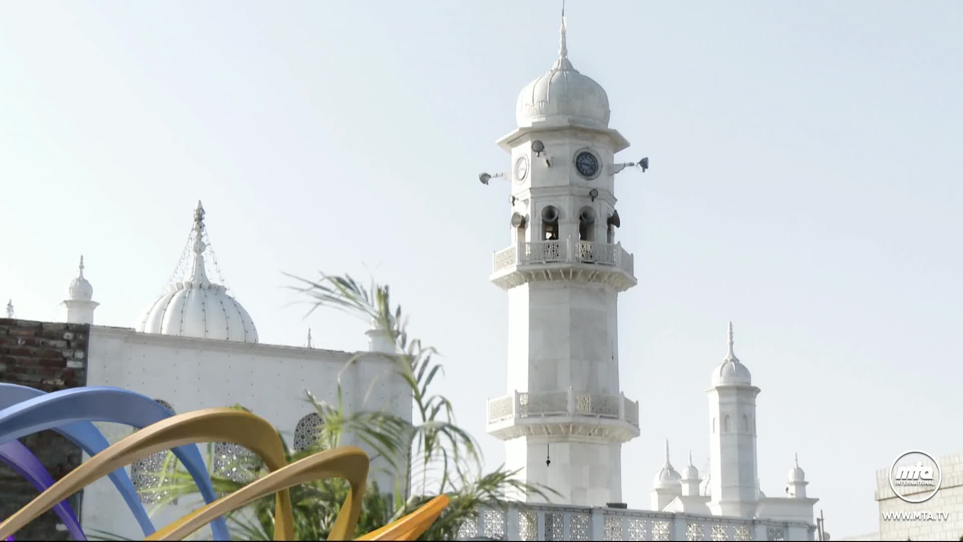 A white minaret rises against a clear sky, its clock tower and speakers visible. The building's ornate details and the MTA1 World logo hint at its significance.

