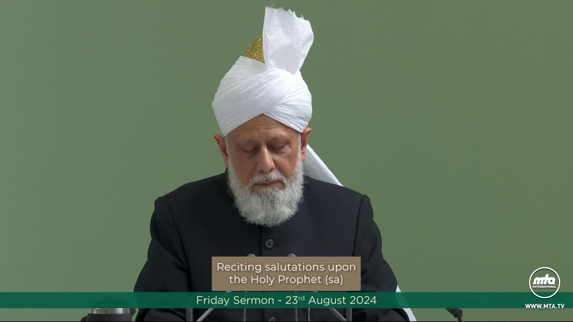 A man wearing a white turban and black suit is speaking, his eyes closed, with a caption below reading "Reciting salutations upon the Holy Prophet (sa)." The broadcast is from MTA1 World, and the date is August 23rd, 2024.
