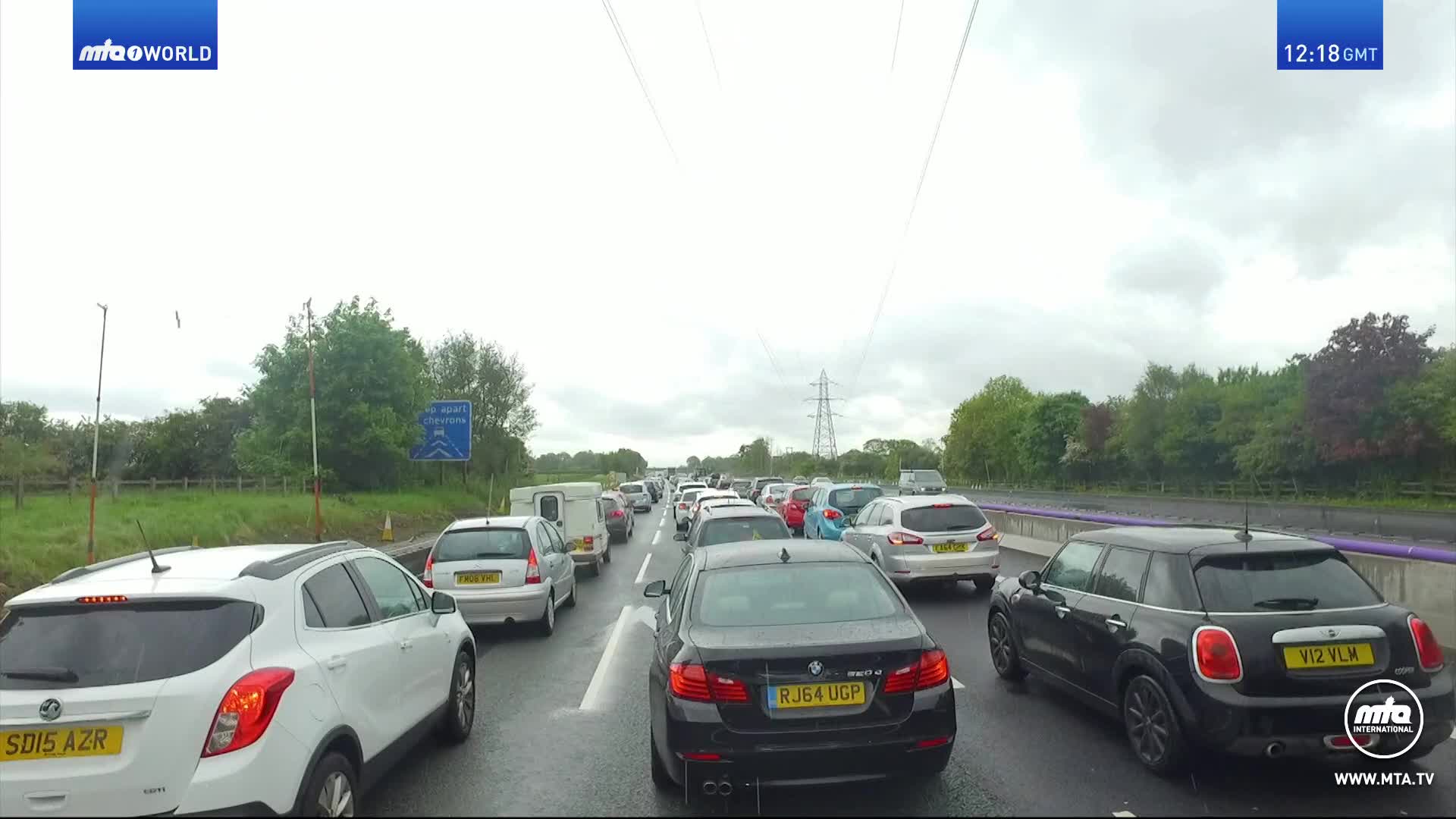 Traffic is at a standstill on a wet UK motorway, with a long line of cars stretching as far as the eye can see. The sky is overcast, and the only movement comes from the occasional windshield wiper.
