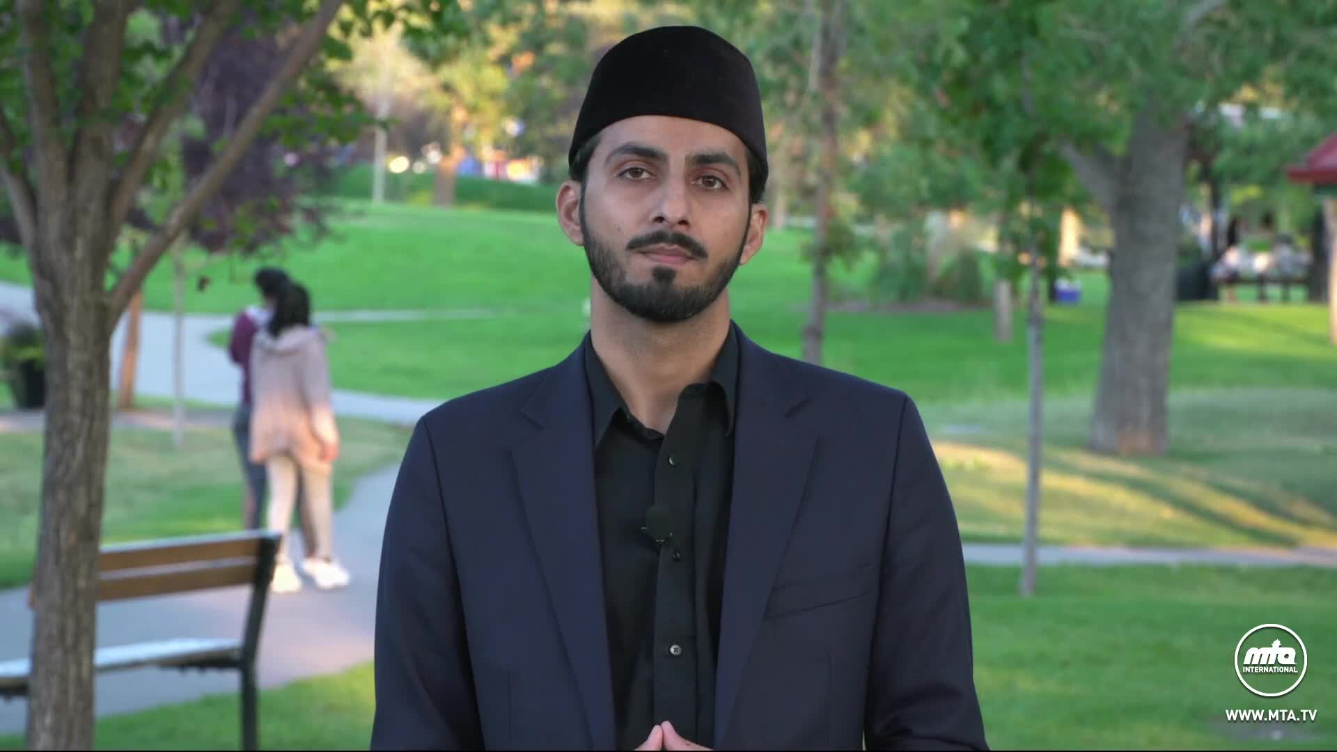 A man in a dark suit and hat is speaking, a small microphone clipped to his lapel. Behind him, a park scene with a bench and two people walking in the distance provides the backdrop.
