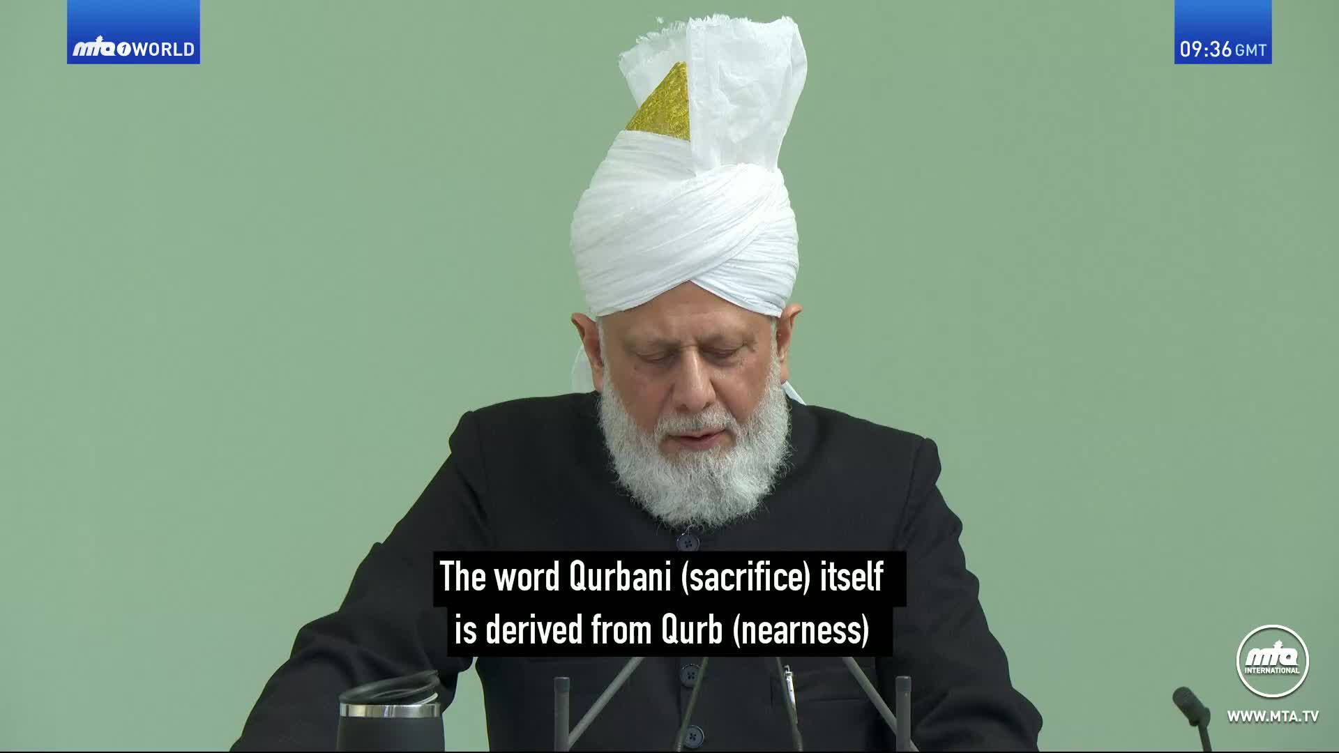 A man with a white turban and beard speaks into a microphone. He is seated behind a table, with the MTA World logo visible in the corner.