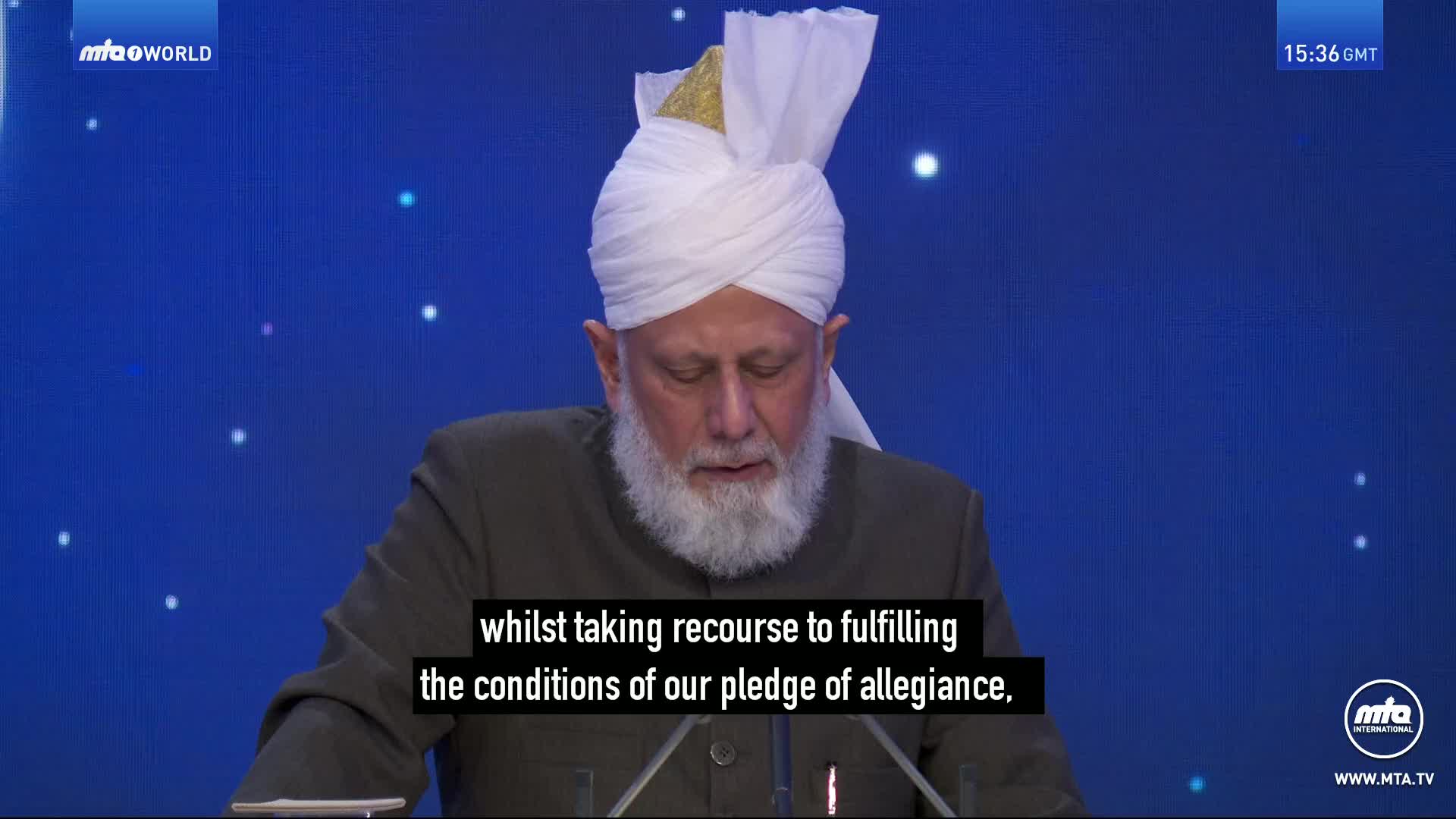 A man in a white turban and dark jacket speaks at a podium. The screen behind him displays a blue background with faint white dots.