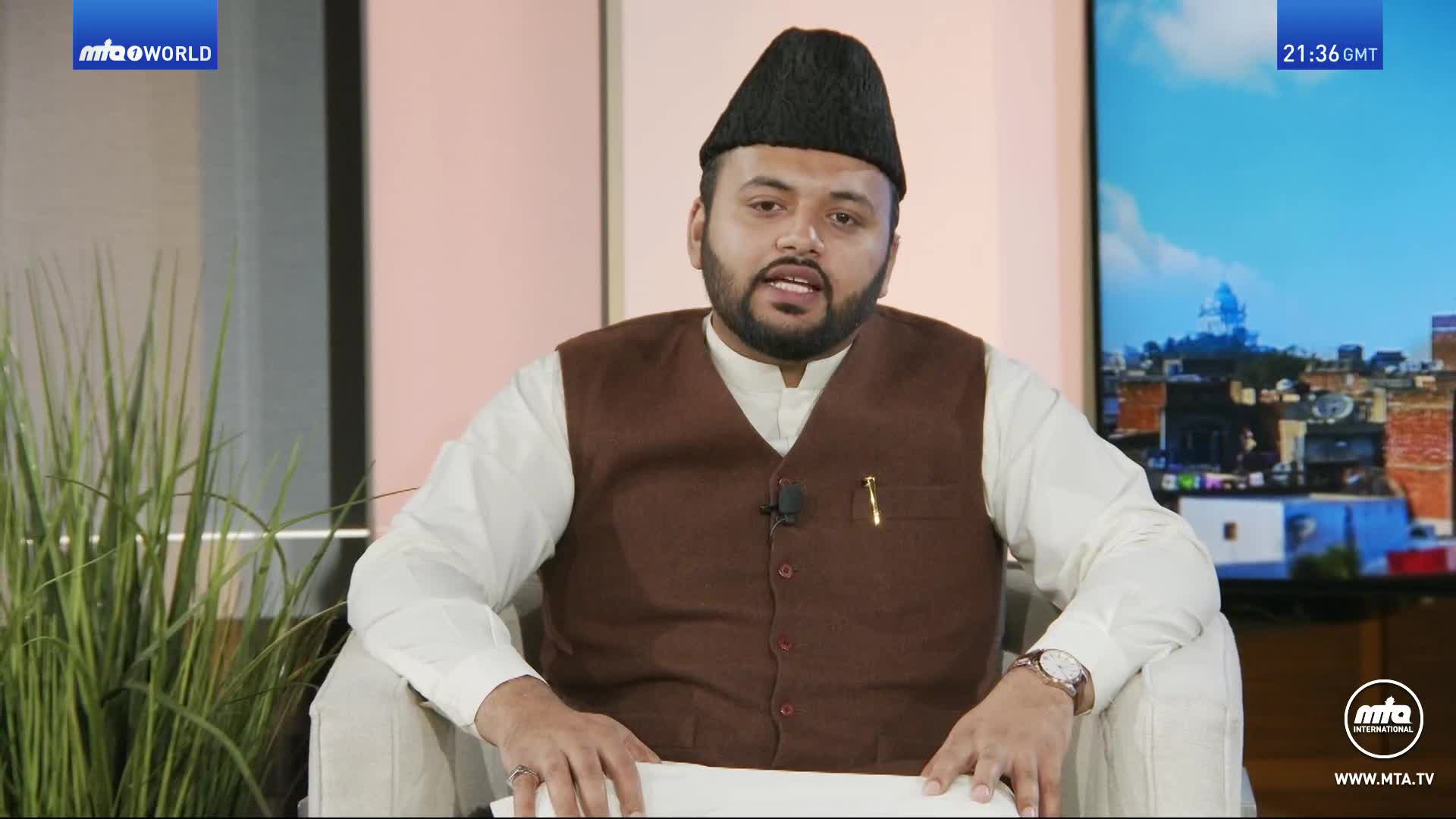 A man in a brown vest and a black cap speaks from a chair, a screen behind him displaying a city skyline and the time 21:36 GMT. A tall plant stands to his left.