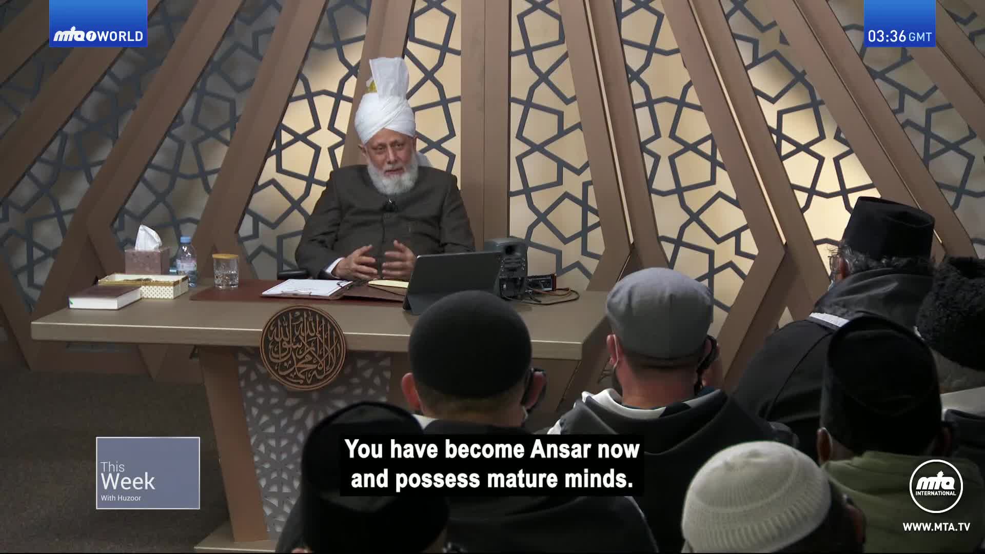 A man in a white turban and traditional clothing speaks from behind a desk, gesturing with his hands. Several people, mostly men wearing skullcaps, listen attentively from chairs arranged in front of him. A man in a white turban and traditional clothing speaks from behind a desk, gesturing with his hands. Several people, mostly men wearing skullcaps, listen attentively from chairs arranged in front of him.