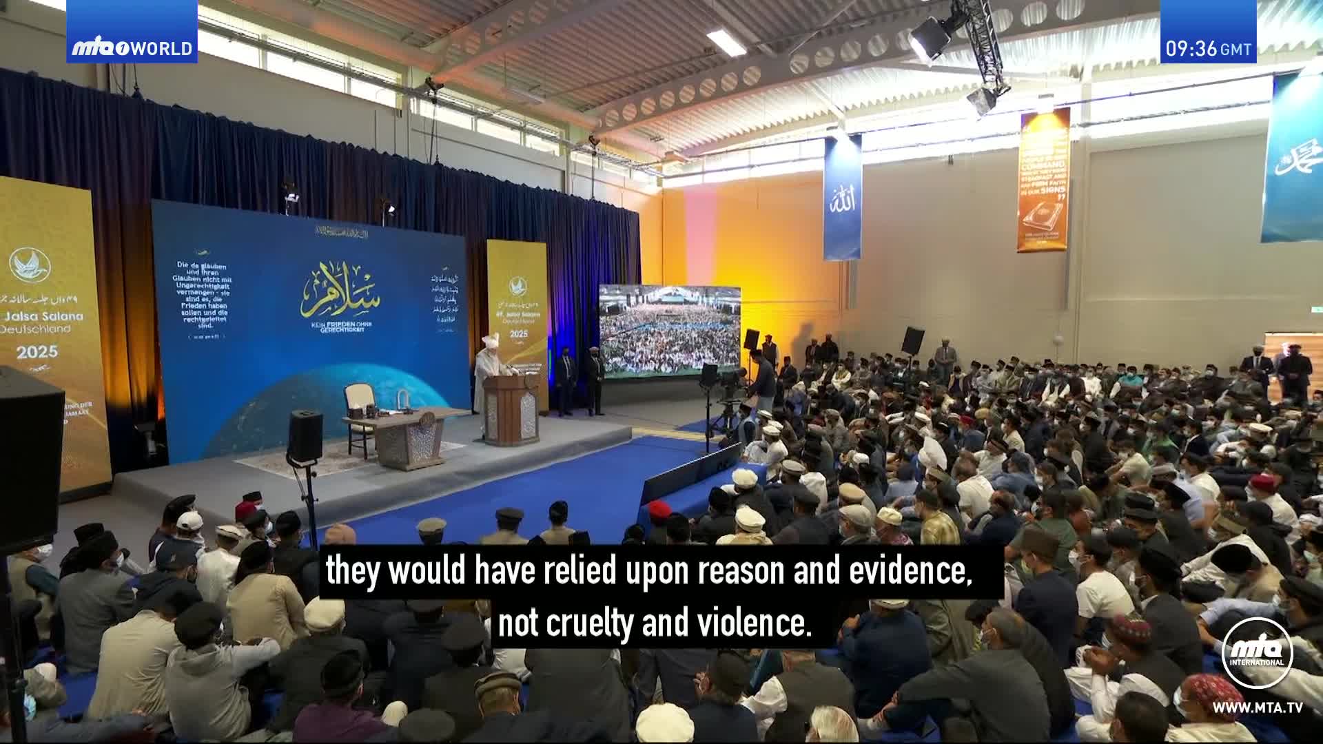 A speaker stands at a podium on a stage in front of a large audience. A screen behind the speaker shows a crowd of people. A speaker stands at a podium on a stage in front of a large audience. A screen behind the speaker shows a crowd of people.