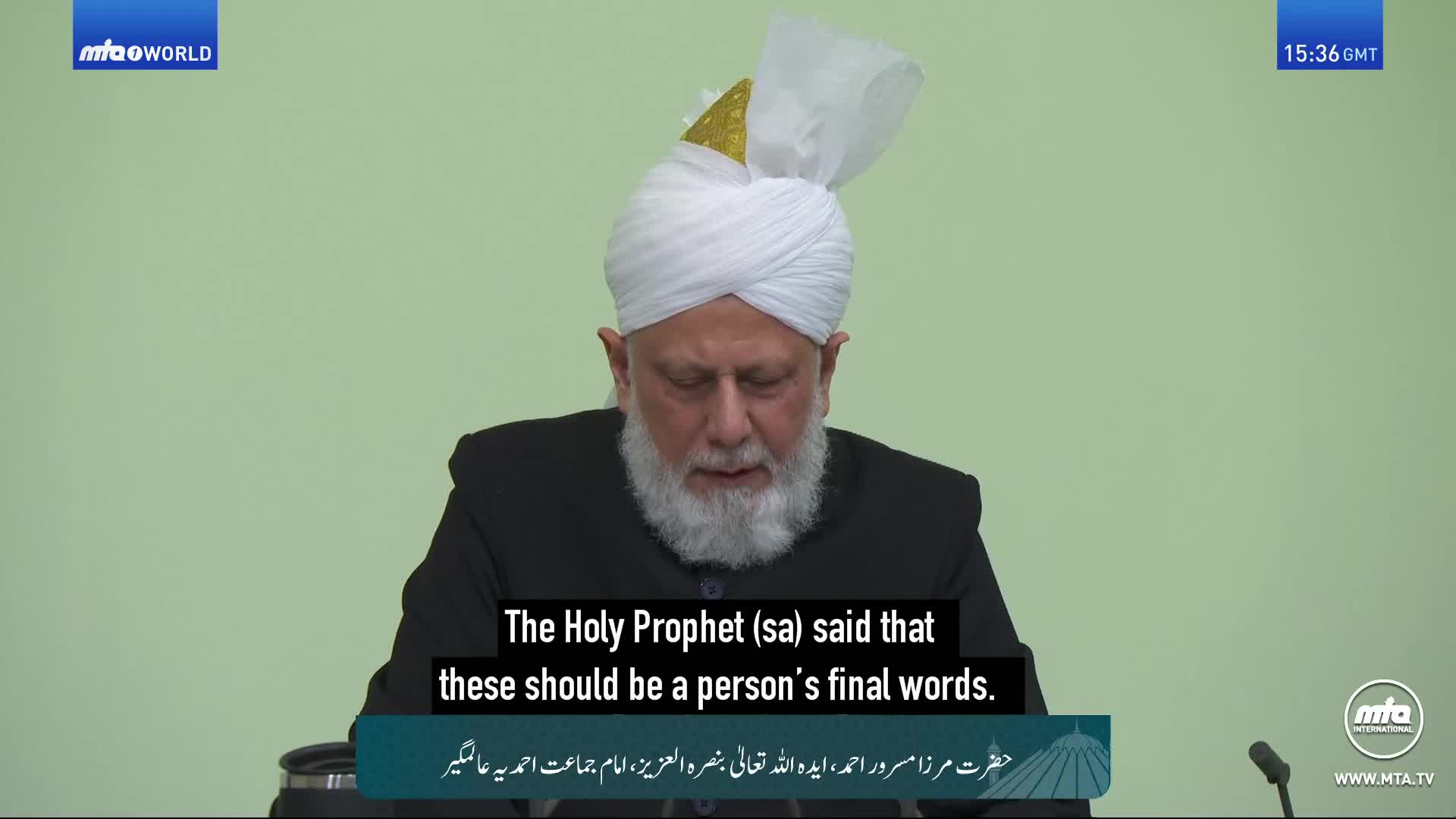 A man in a white turban and dark jacket speaks, his gaze lowered. The screen displays subtitles in English and Arabic, indicating a broadcast from the United Kingdom. A man in a white turban and dark jacket speaks, his gaze lowered. The screen displays subtitles in English and Arabic, indicating a broadcast from the United Kingdom.