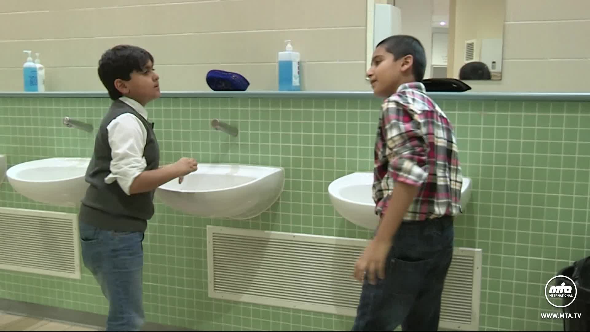 Two boys stand by sinks in a washroom. One boy, in a grey vest over a white shirt, gestures with his hand. The other boy, in a red plaid shirt, faces him. Two boys stand by sinks in a washroom. One boy, in a grey vest over a white shirt, gestures with his hand. The other boy, in a red plaid shirt, faces him.