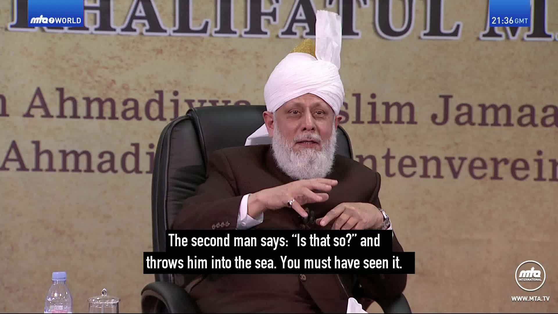 A man in a white turban and brown jacket gestures with his hands as he speaks. The screen displays text about a man being thrown into the sea. A man in a white turban and brown jacket gestures with his hands as he speaks. The screen displays text about a man being thrown into the sea.