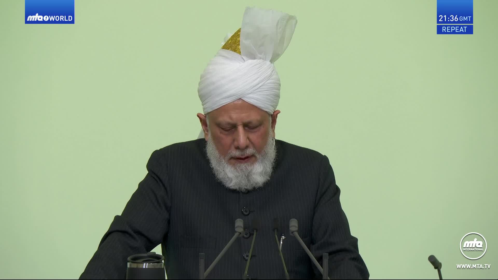 A man in a white turban and dark suit stands at a podium, his eyes closed. The screen displays "mtaoworld" and "21:36 GMT REPEAT." A man in a white turban and dark suit stands at a podium, his eyes closed. The screen displays "mtaoworld" and "21:36 GMT REPEAT."