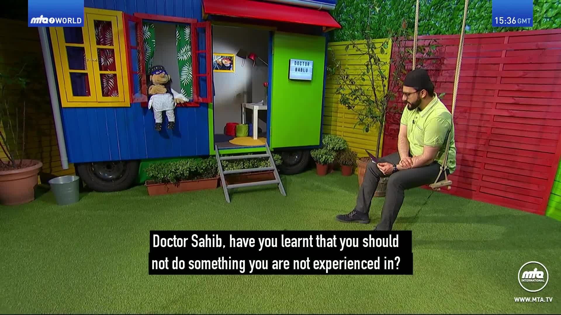 A puppet sits in the window of a brightly colored caravan. A man in a light green shirt is seated on a swing, looking at a tablet.