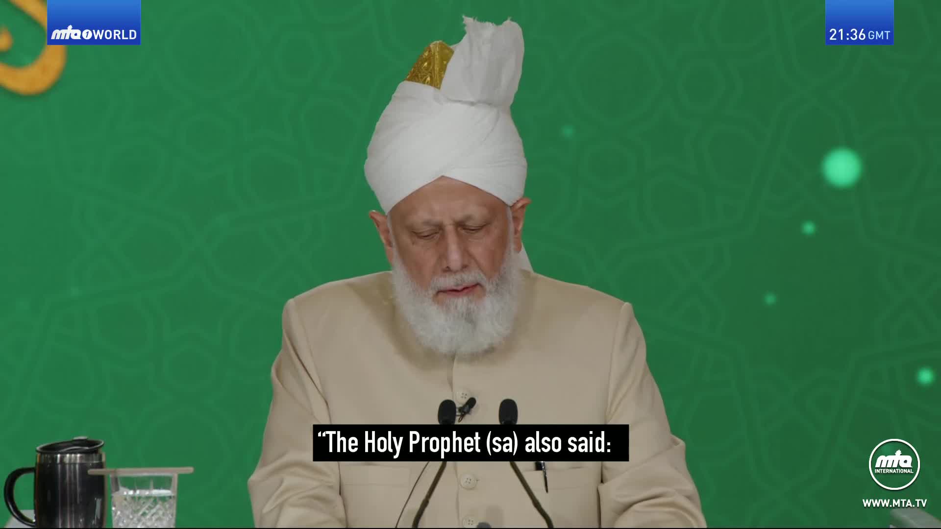 The man in the white turban and beige jacket looks down as he speaks. A caption below him reads, "The Holy Prophet (sa) also said:".