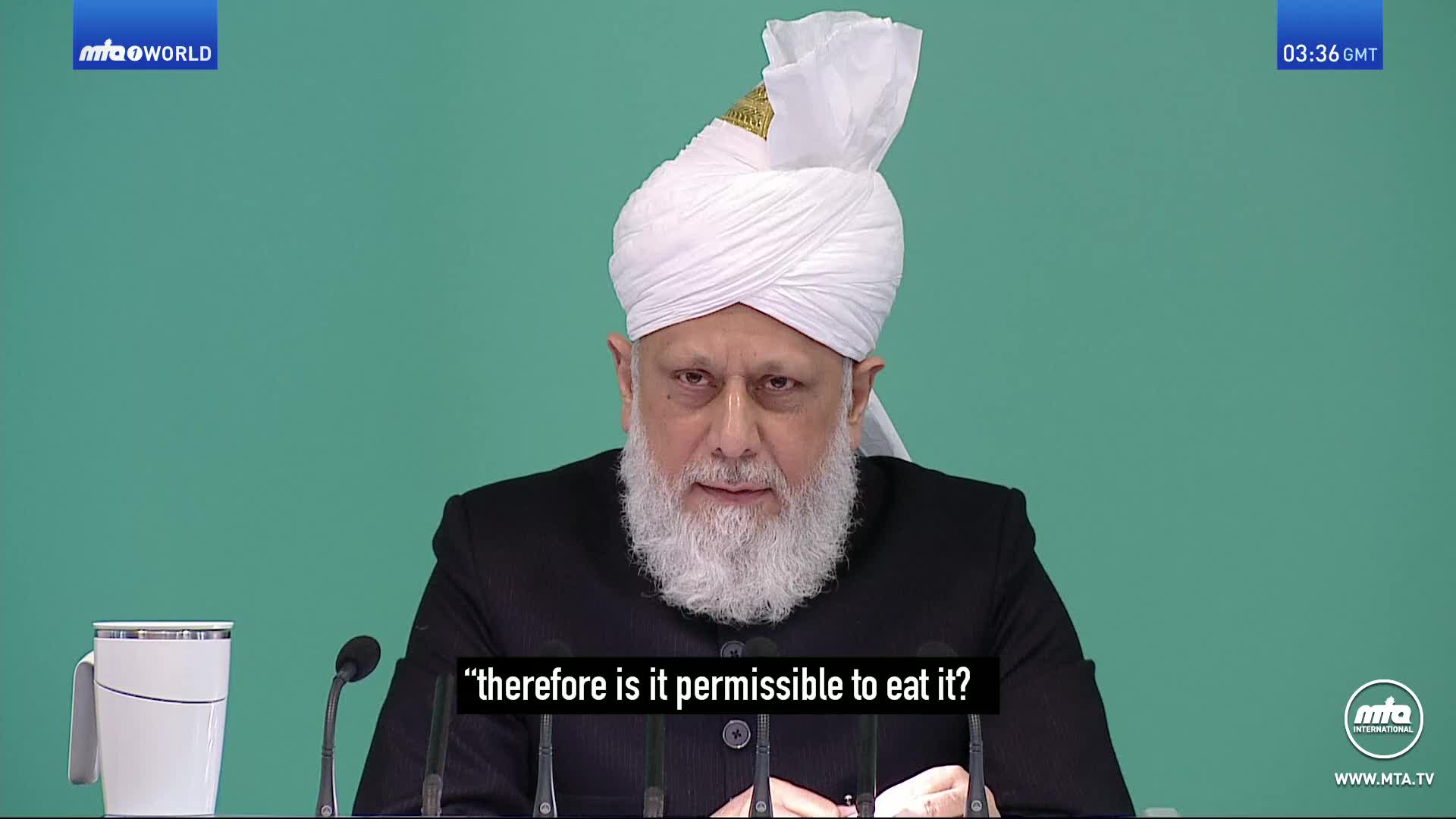 A man with a white turban and beard speaks, his words appearing on screen. A tall white mug sits to his left.