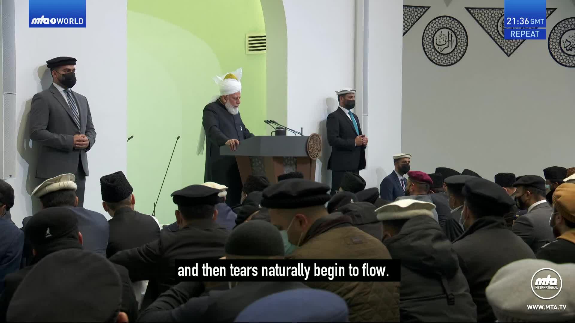 A man in a white turban stands at a podium, addressing a seated congregation. Two men in dark suits stand to his left and right, observing the crowd.