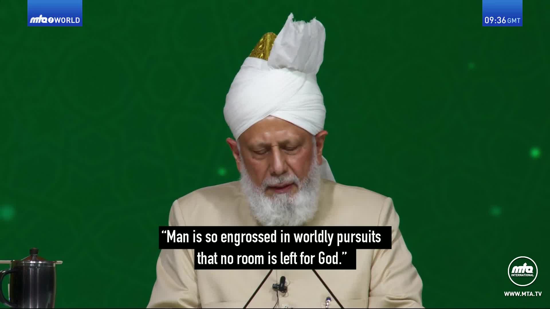 A man in a white turban and cream jacket speaks from a stage with a green backdrop. A quote appears on screen: "Man is so engrossed in worldly pursuits that no room is left for God."