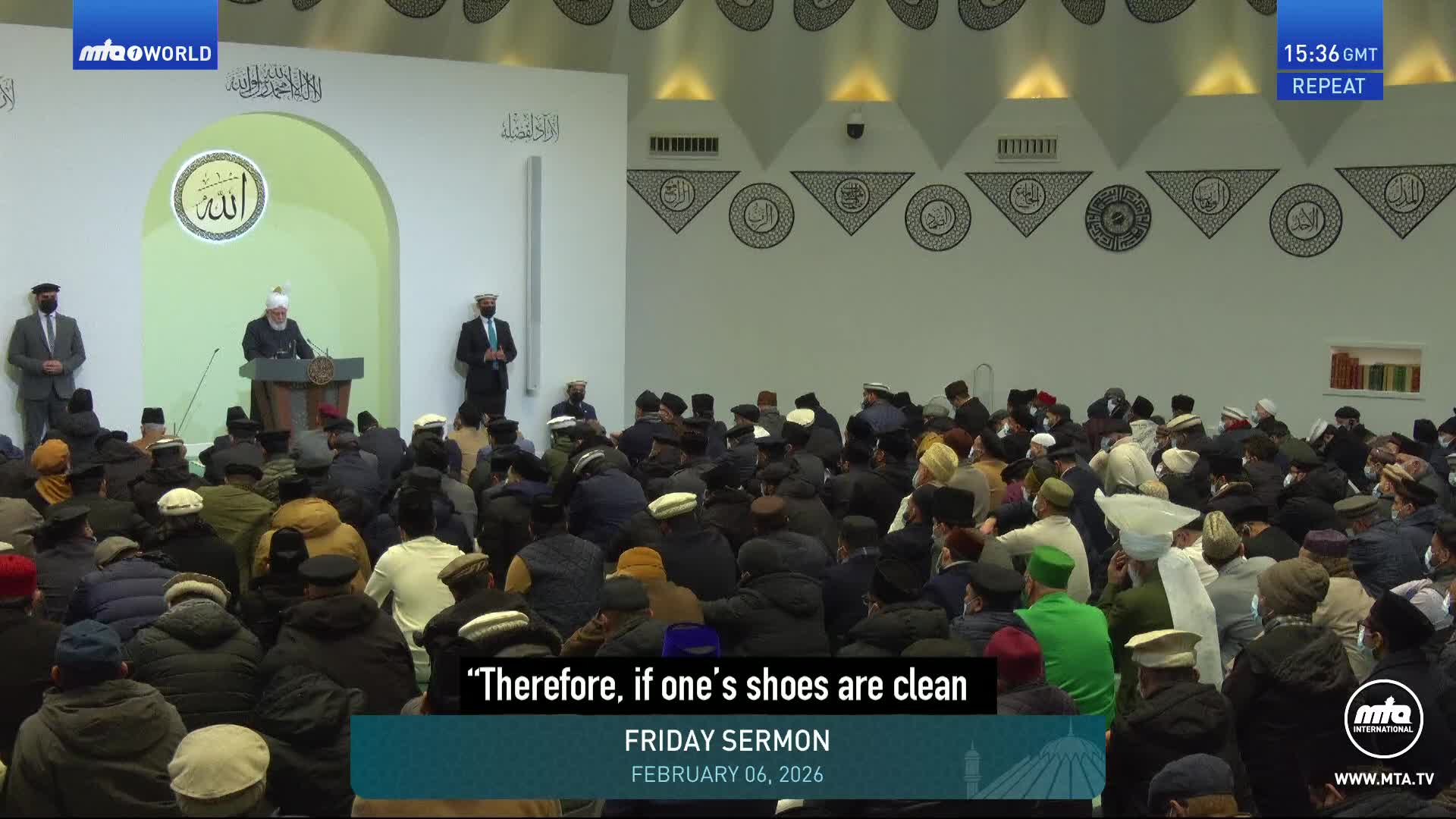 A man in a white turban stands at a podium, addressing a large congregation seated before him. The room is filled with men wearing various hats and caps, listening intently to the Friday sermon.