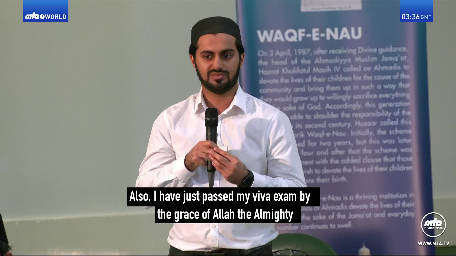 A man in a white shirt and black cap holds a microphone, speaking into it. Behind him, a banner details the Waqf-e-Nau scheme.