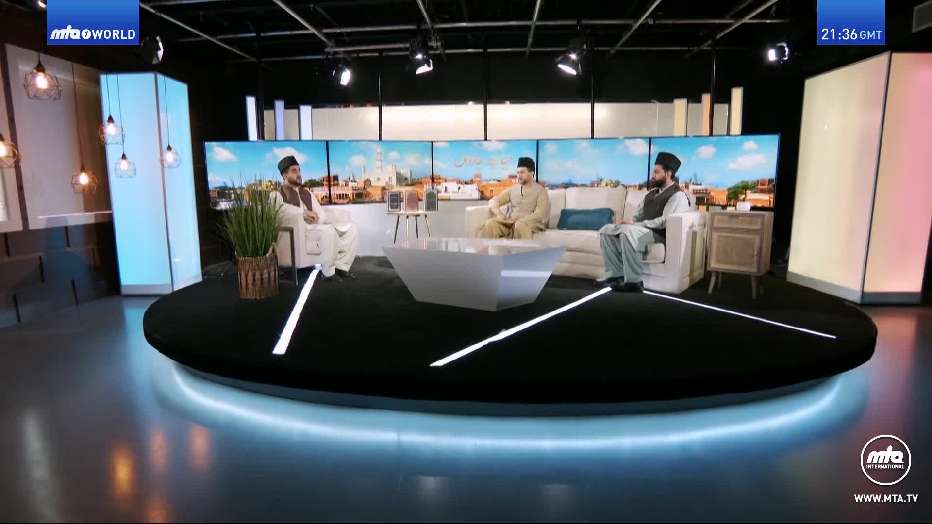 Three men in traditional Islamic attire are seated on a modern set, facing each other across a white coffee table. A large screen behind them displays a cityscape under a bright sky, suggesting a broadcast from MTA World.