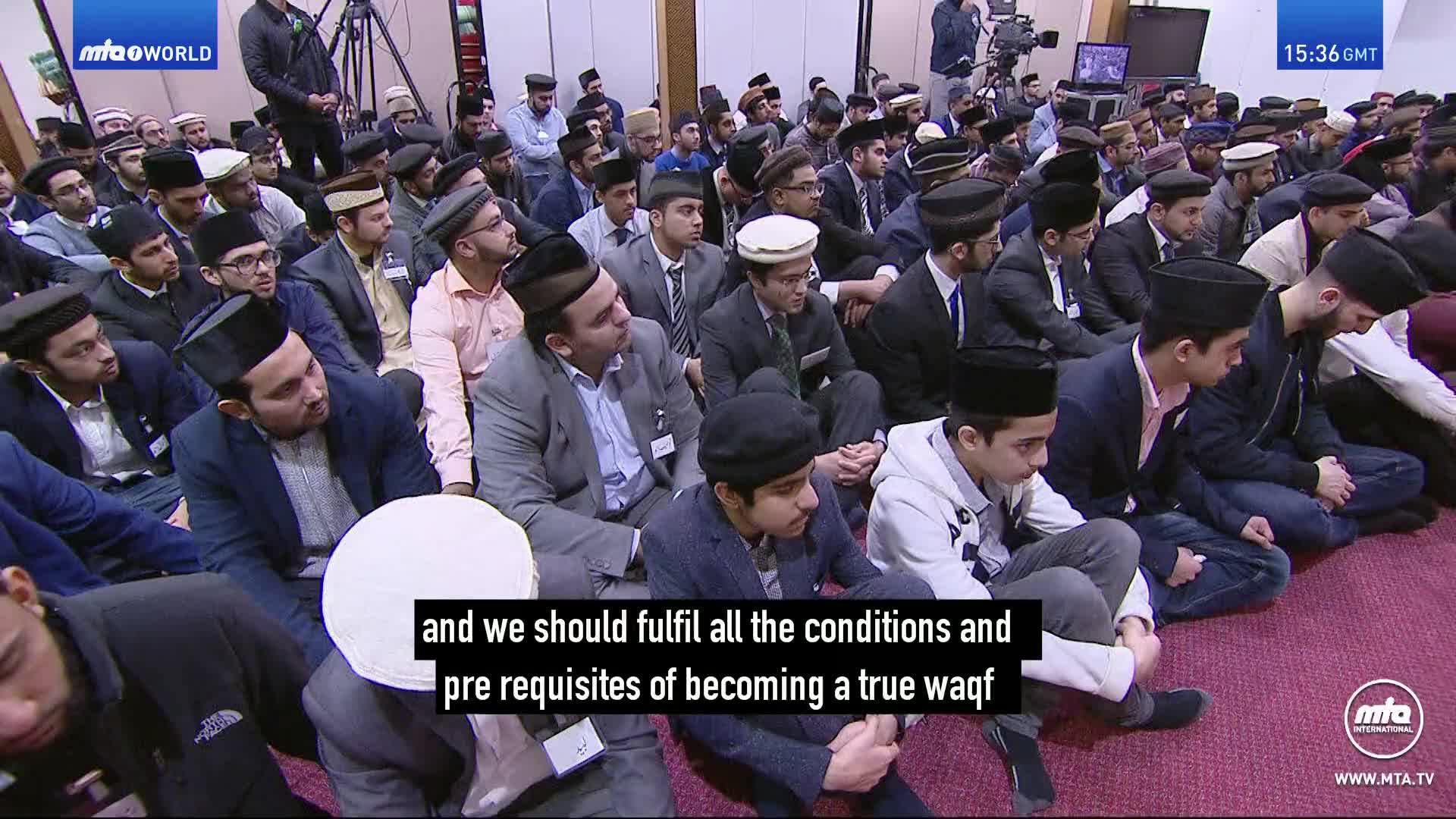 A large group of men and boys, many wearing traditional Islamic caps, sit on a carpeted floor. A speaker addresses them from a raised platform, while cameras capture the event.