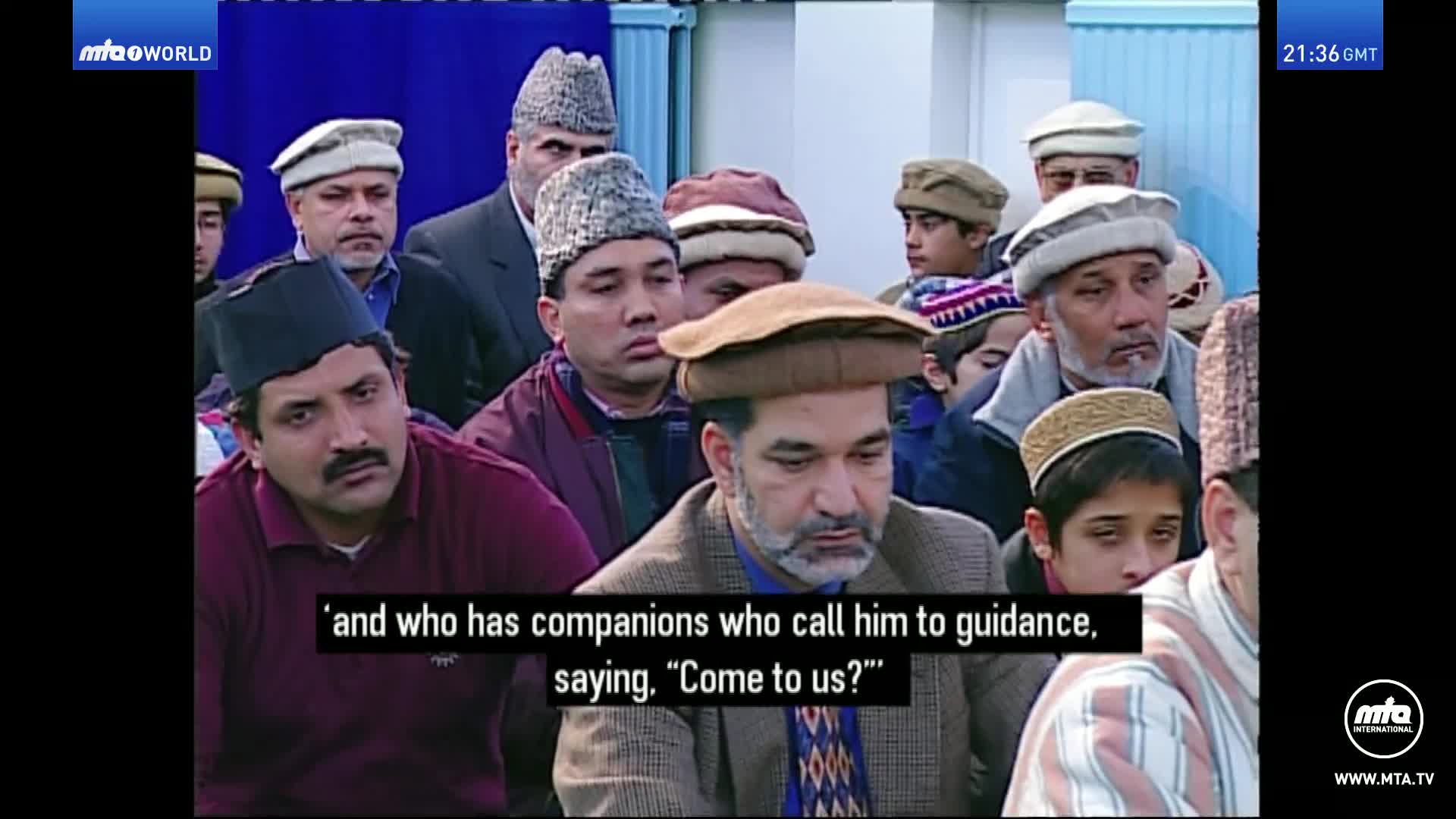 A group of men and boys, many wearing traditional caps, are gathered, listening intently. A man in the foreground with a beard and a patterned tie looks down as words appear on screen.