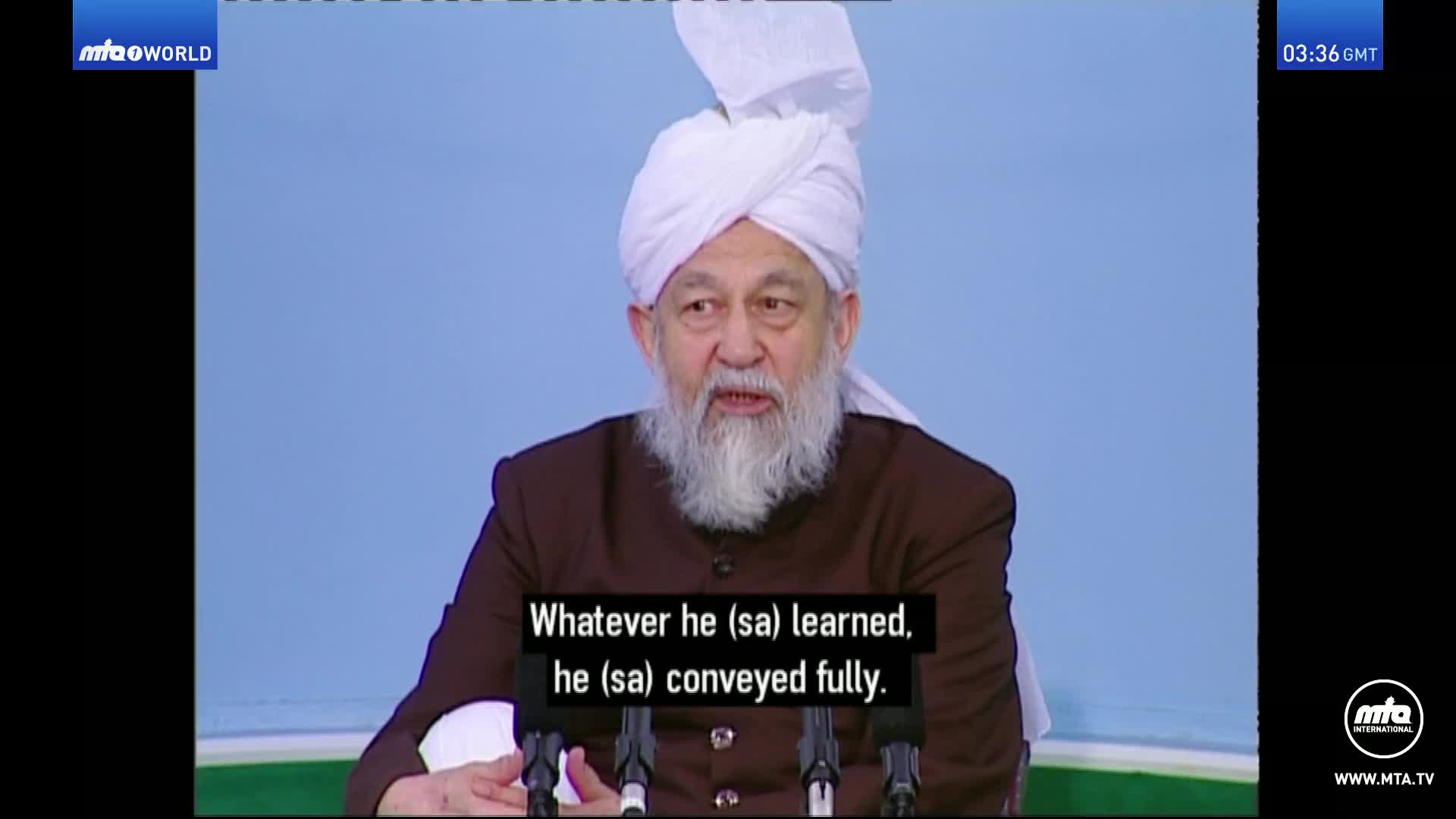 A man in a white turban and brown jacket speaks into a microphone. The MTA World logo appears in the top left corner.