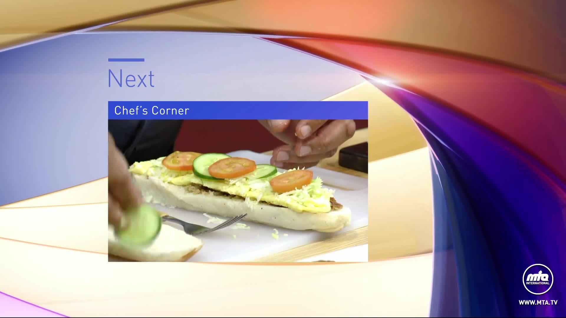 The next segment, "Chef's Corner," is about to begin. A hand places a slice of cucumber onto a long sandwich topped with lettuce, tomato, and egg.