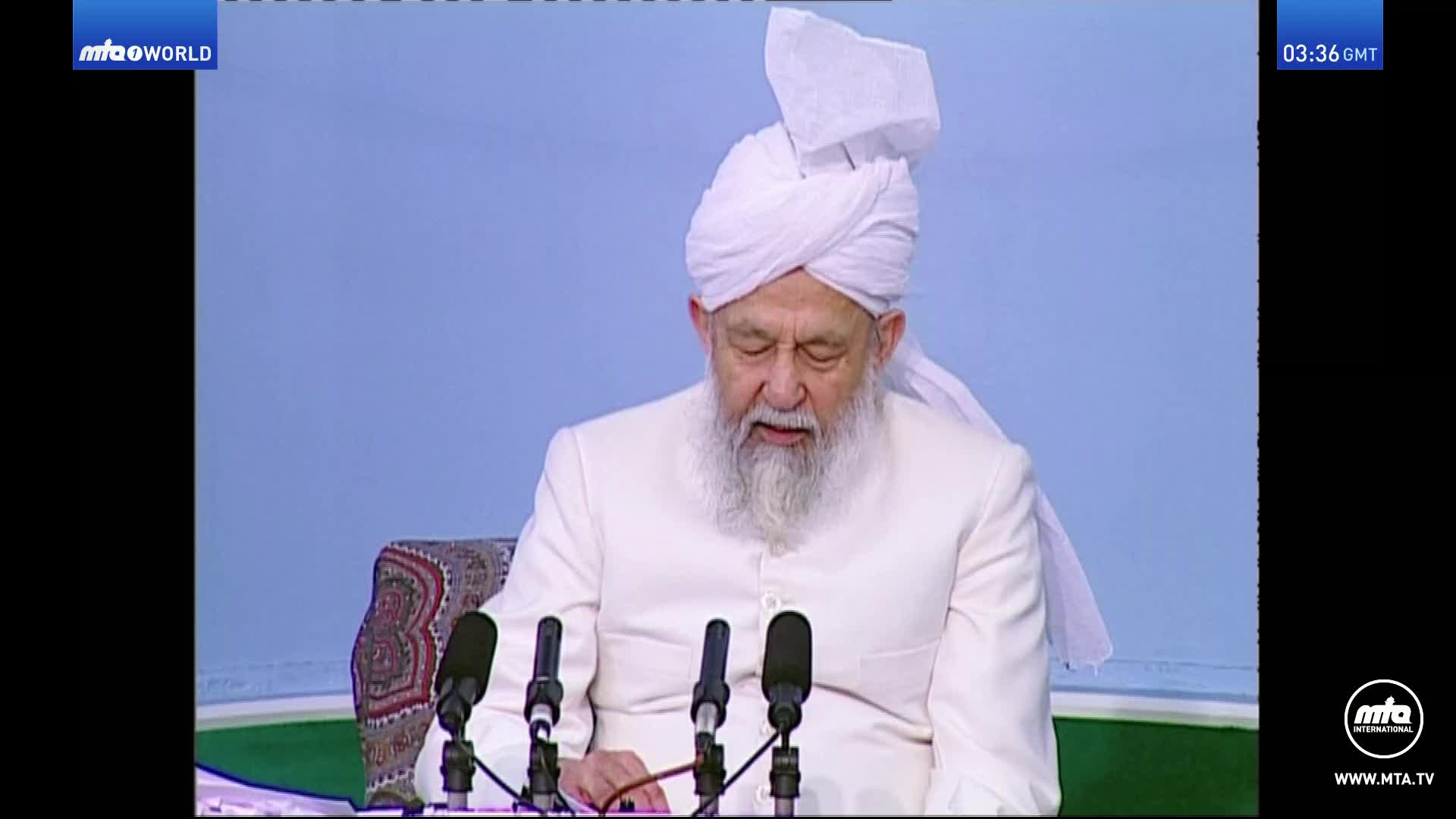 An elder man with a white turban and beard speaks into three microphones. The broadcast shows "mtaowORLD" in the upper left corner and "03:36 GMT" in the upper right.
