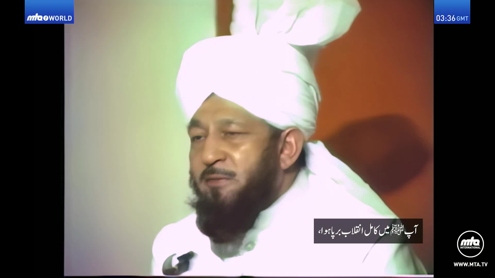 A man in a white turban and robes speaks, his beard framing his face. The MTA World logo appears in the corner, and text in Arabic script is overlaid on the screen.