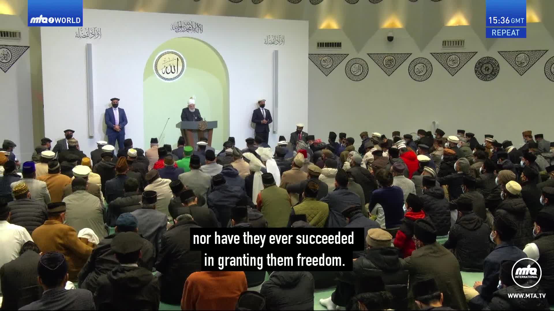 A man in a white turban stands at a podium, addressing a large gathering of men seated on the floor. Another man in a suit stands to the left of the podium, facing the audience.