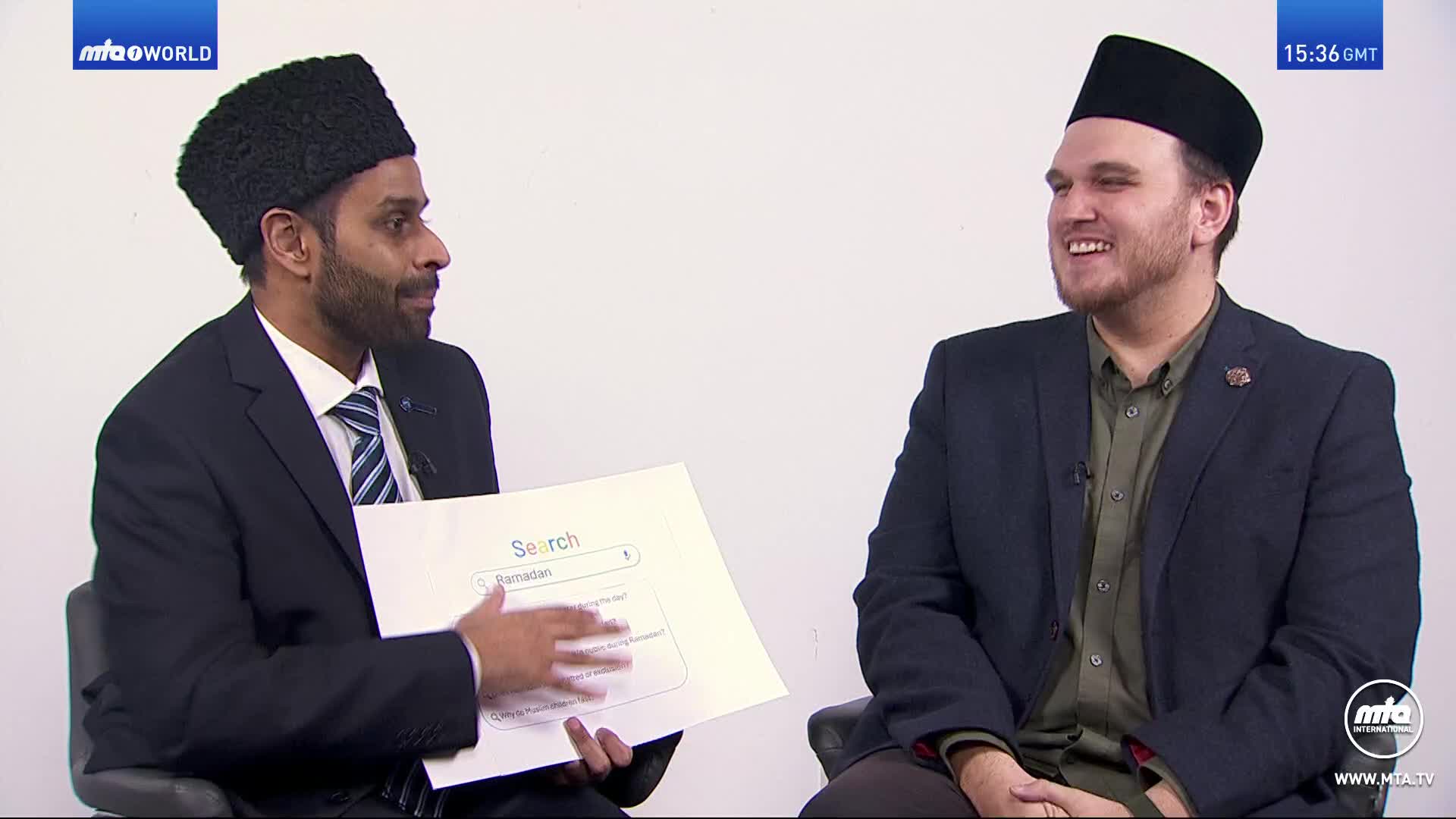A man in a black karakul hat gestures towards a printout of a search engine result page. He's speaking to another man in a dark suit and a black kufi, who is smiling.
