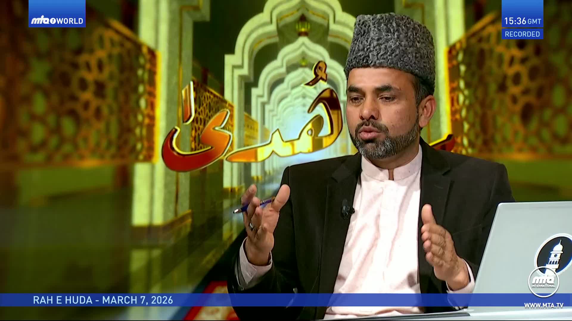 A man in a black suit and a patterned cap speaks with a pen in his hand. Behind him, golden Arabic script glows against a backdrop of ornate arches.