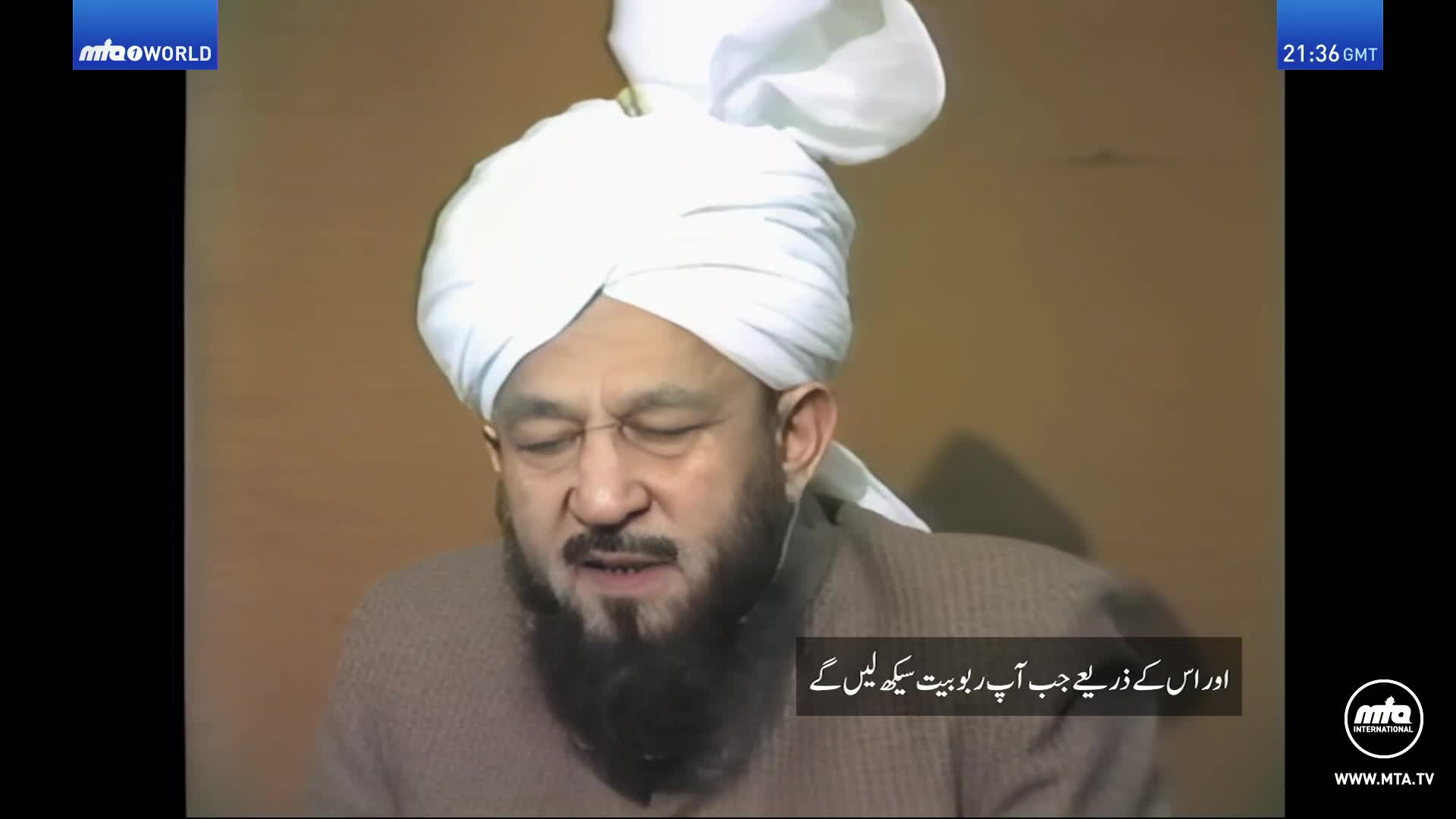 A man in a white turban speaks, his eyes closed. Text in Arabic appears on the screen.