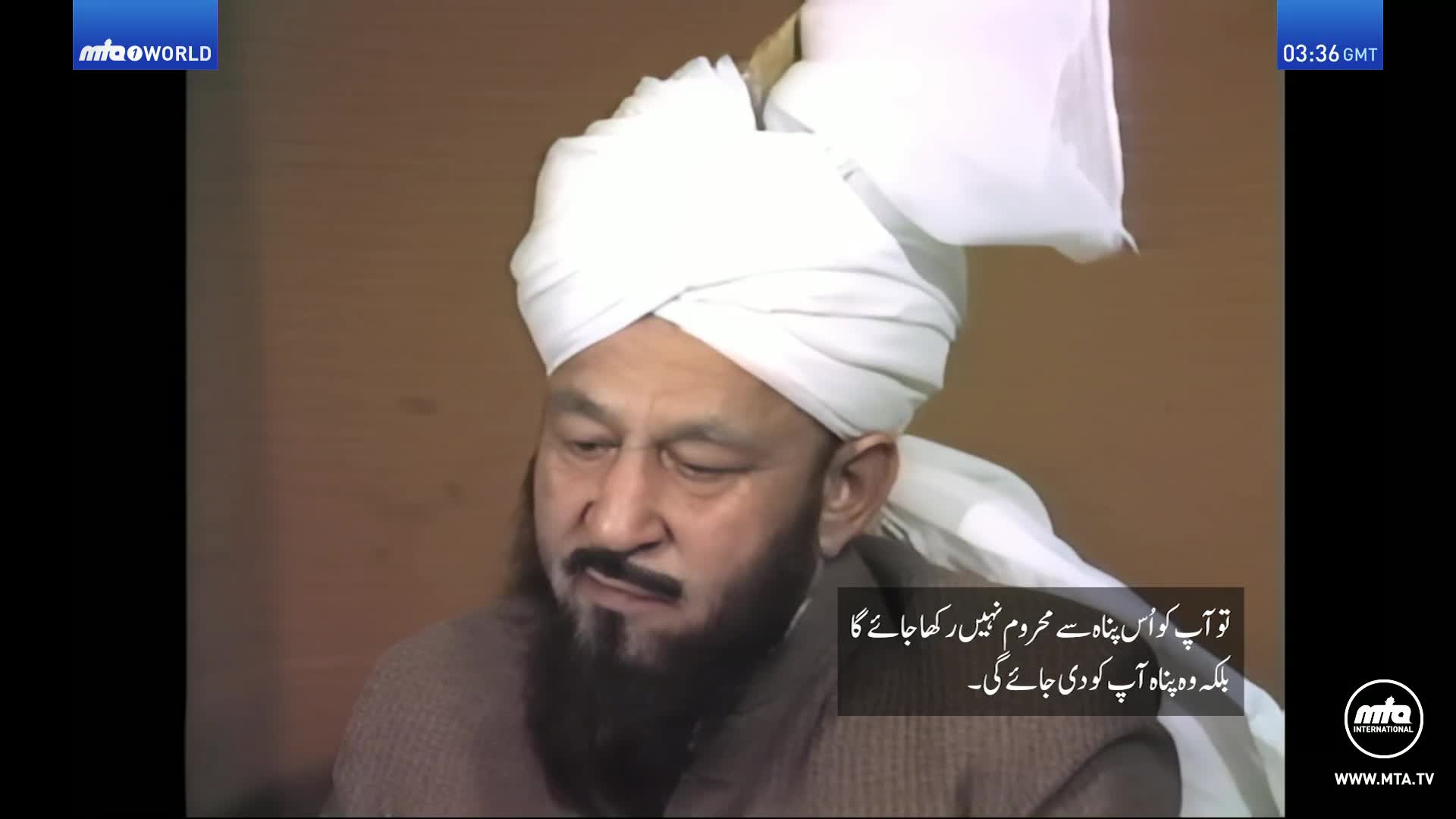 A man with a long beard and a white turban is speaking. Arabic text overlays the bottom right of the screen.
